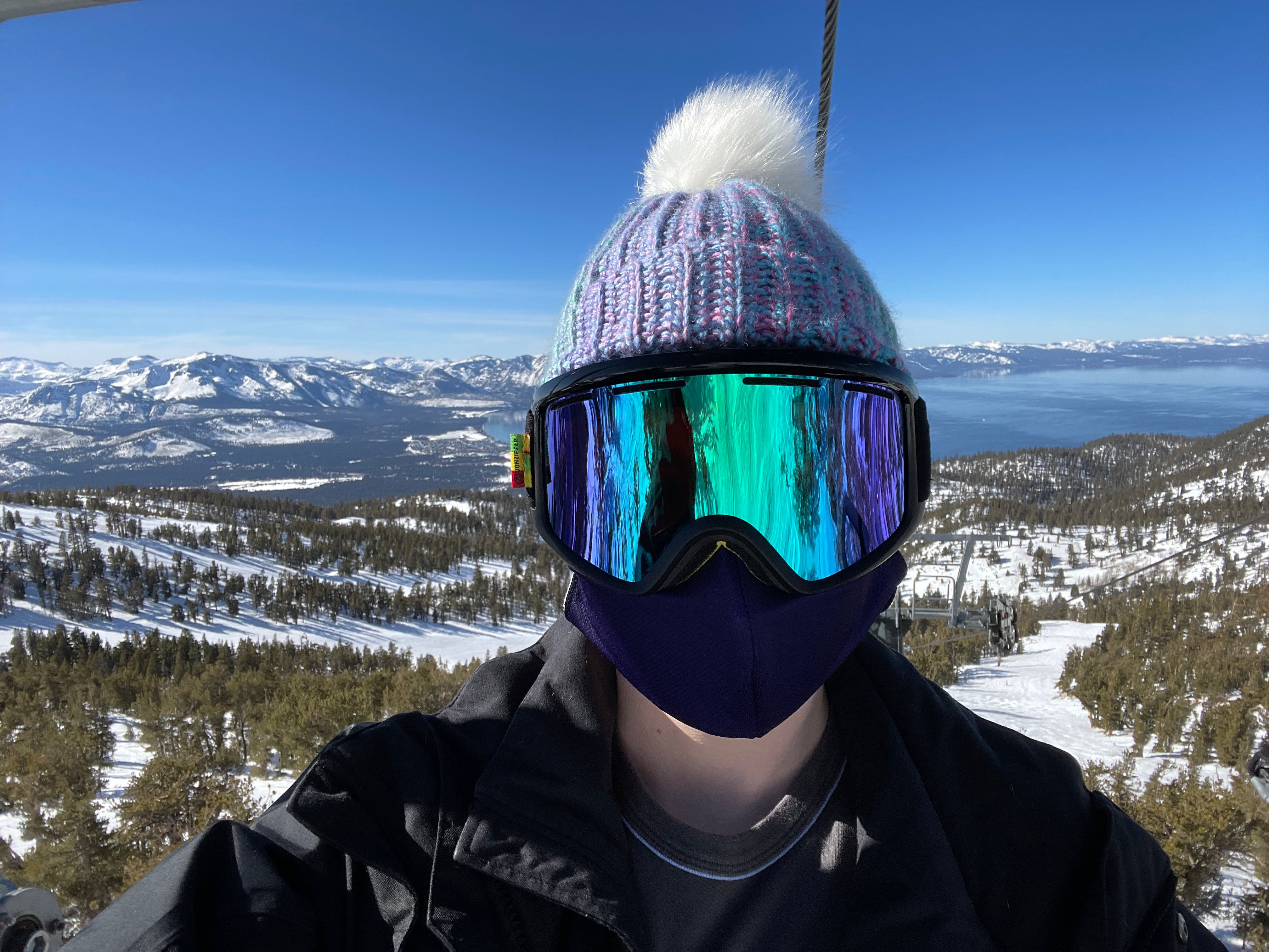 A man in a blue hat and ski googles on a chairlift, with mountains and a lake in the background.