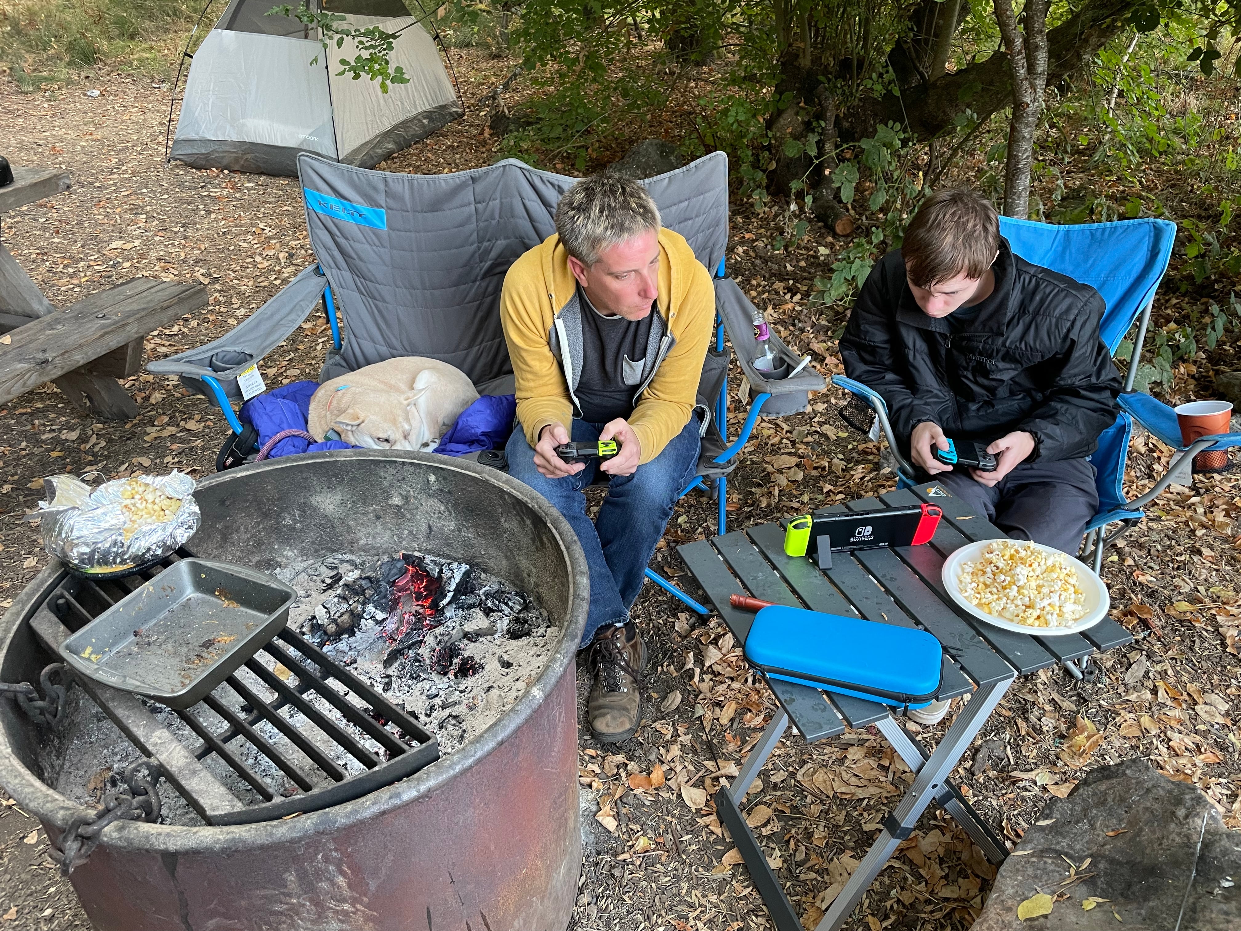 Two men playing videogames while sitting in camping chairs. A dog is also asleep in a camping chair.