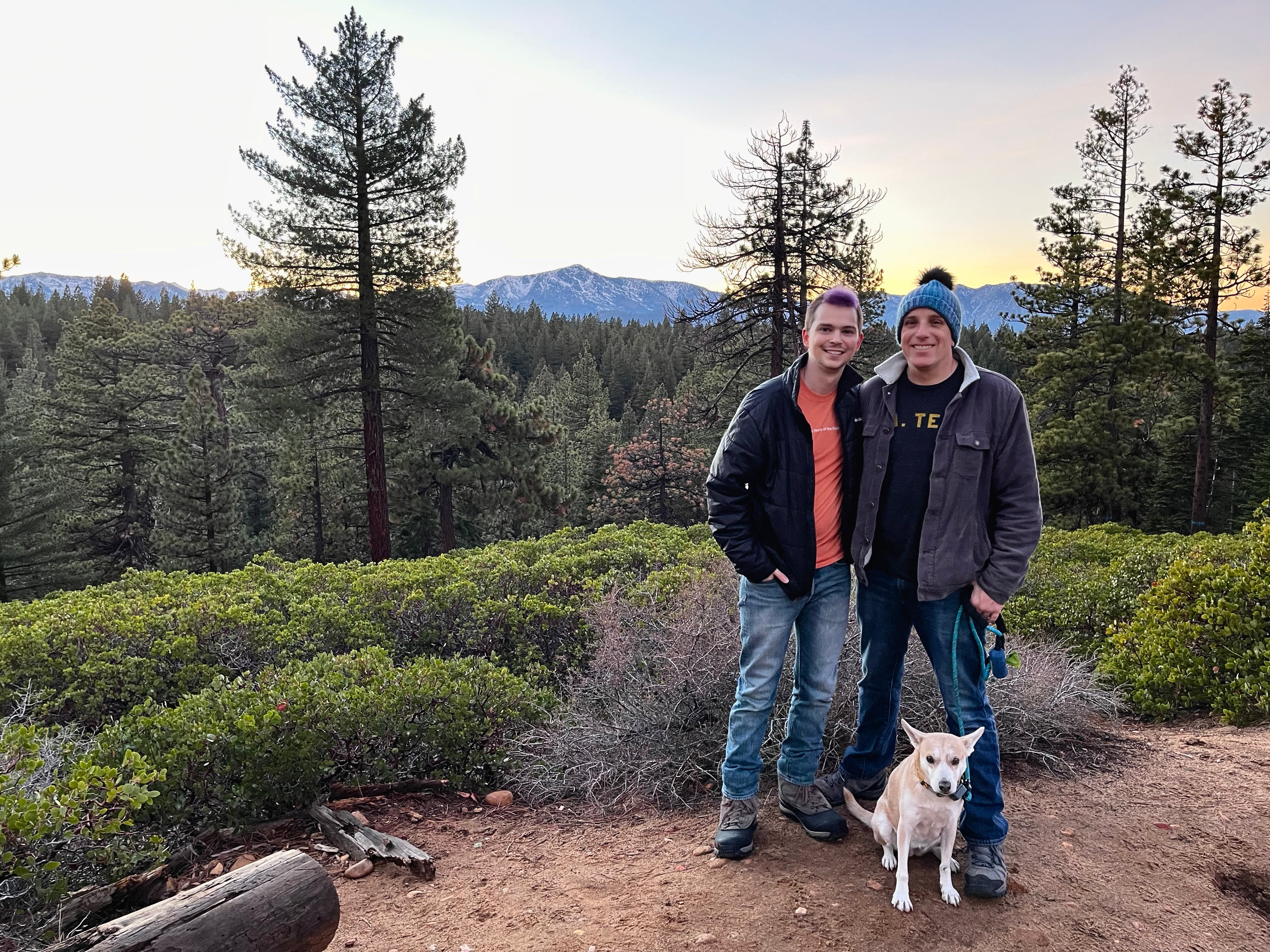Two men and a dog posing for a photo in front of mountains and trees.