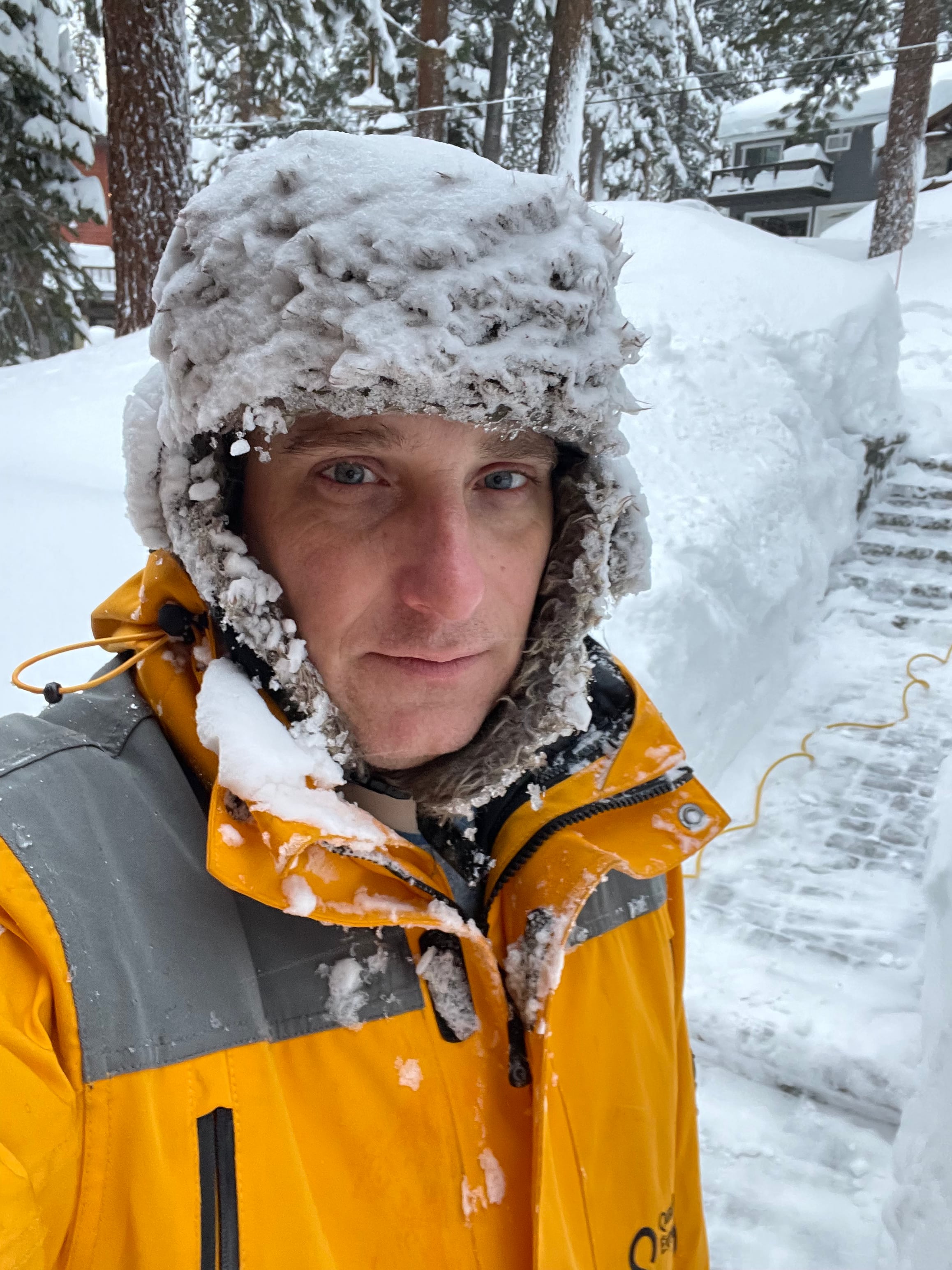 A man smiling, his hat is crusted in snow and ice from clearing snow.