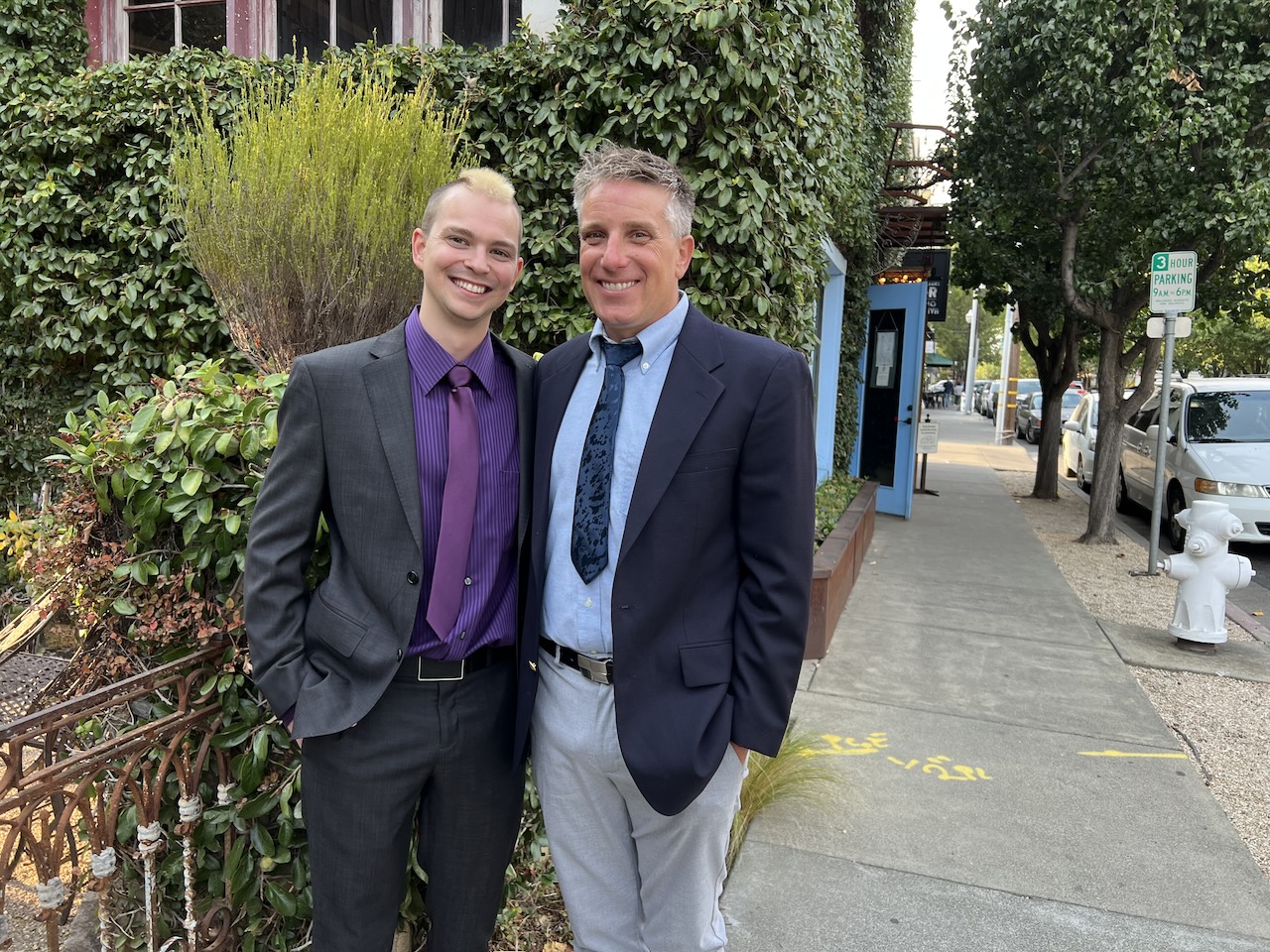 Myke and James standing outside of a brick building and holding drinks.