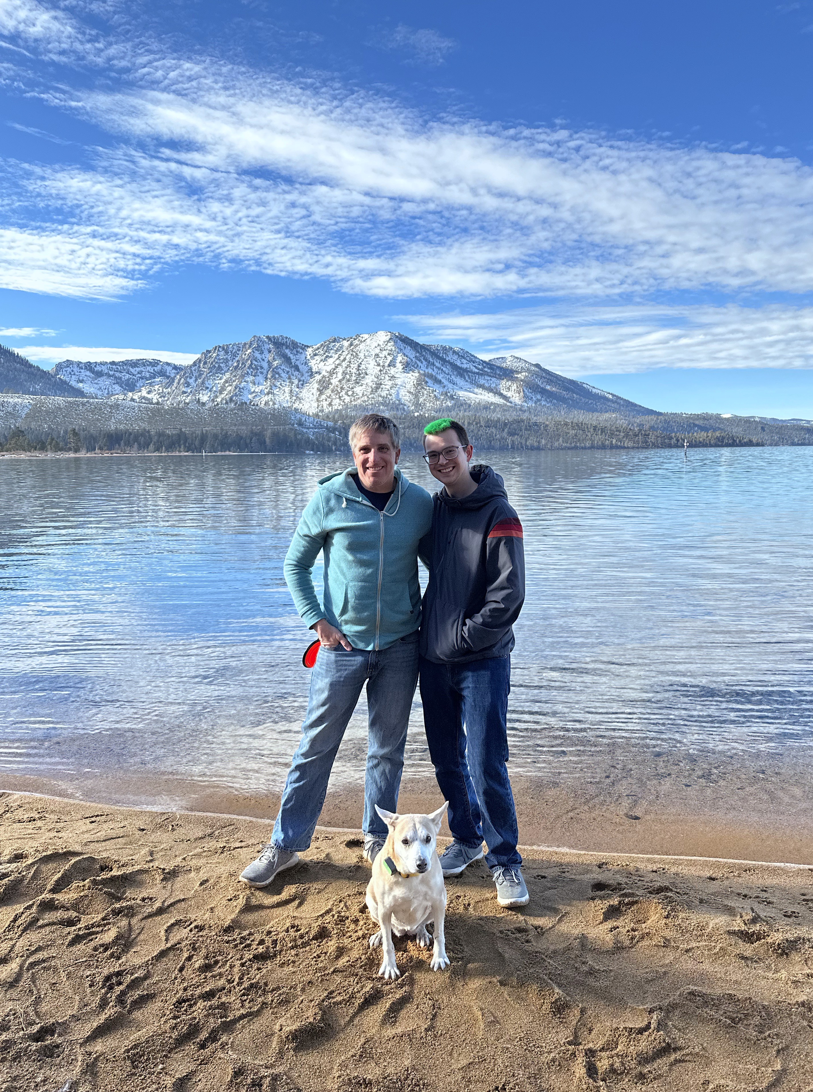 Two people and a medium sized dog posing for a photo on the shore of a lake. A large mountain is visible in the background.