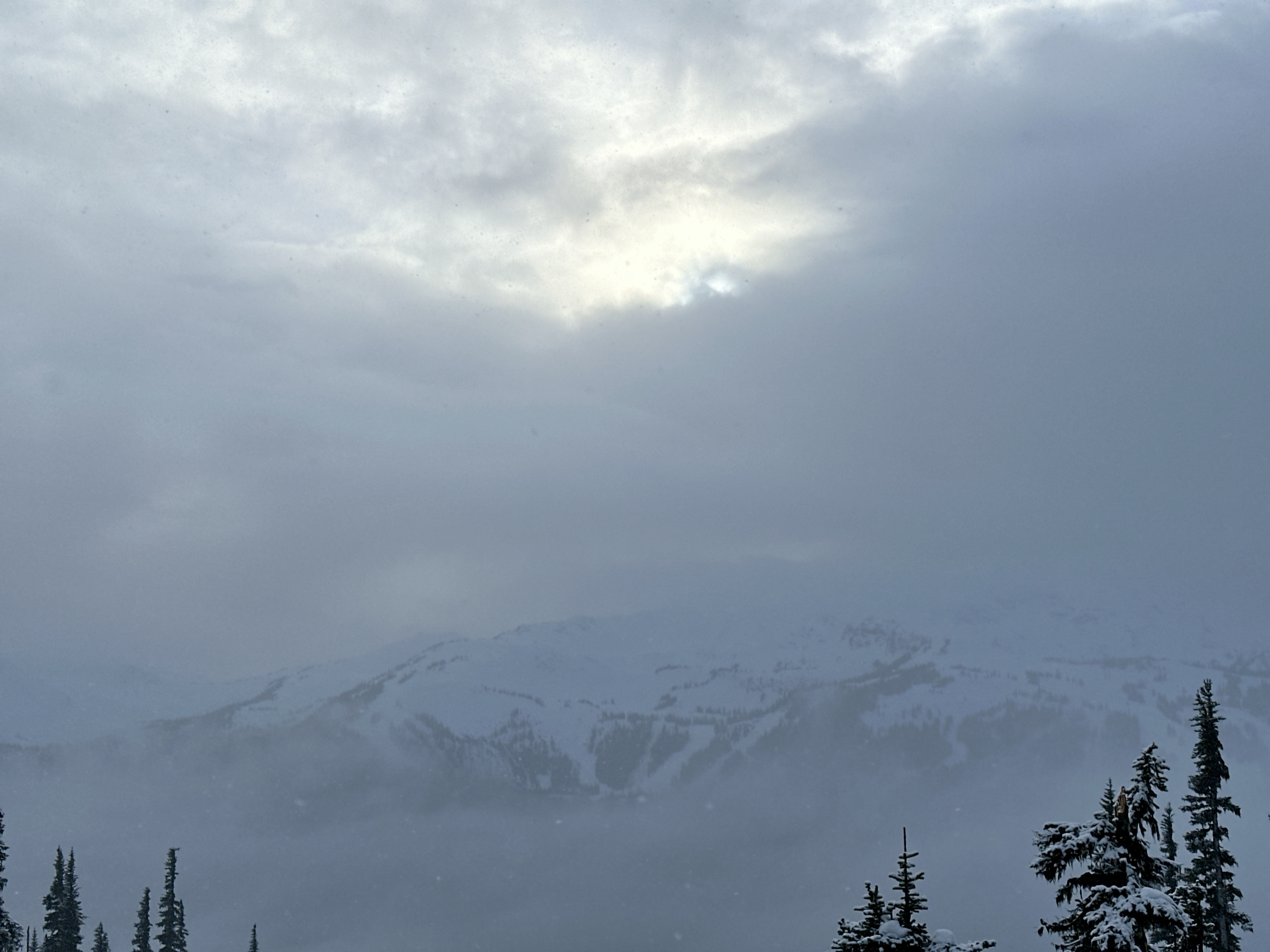 A wide, snow-covered mountain top, only faintly visible thorugh clouds and fog.