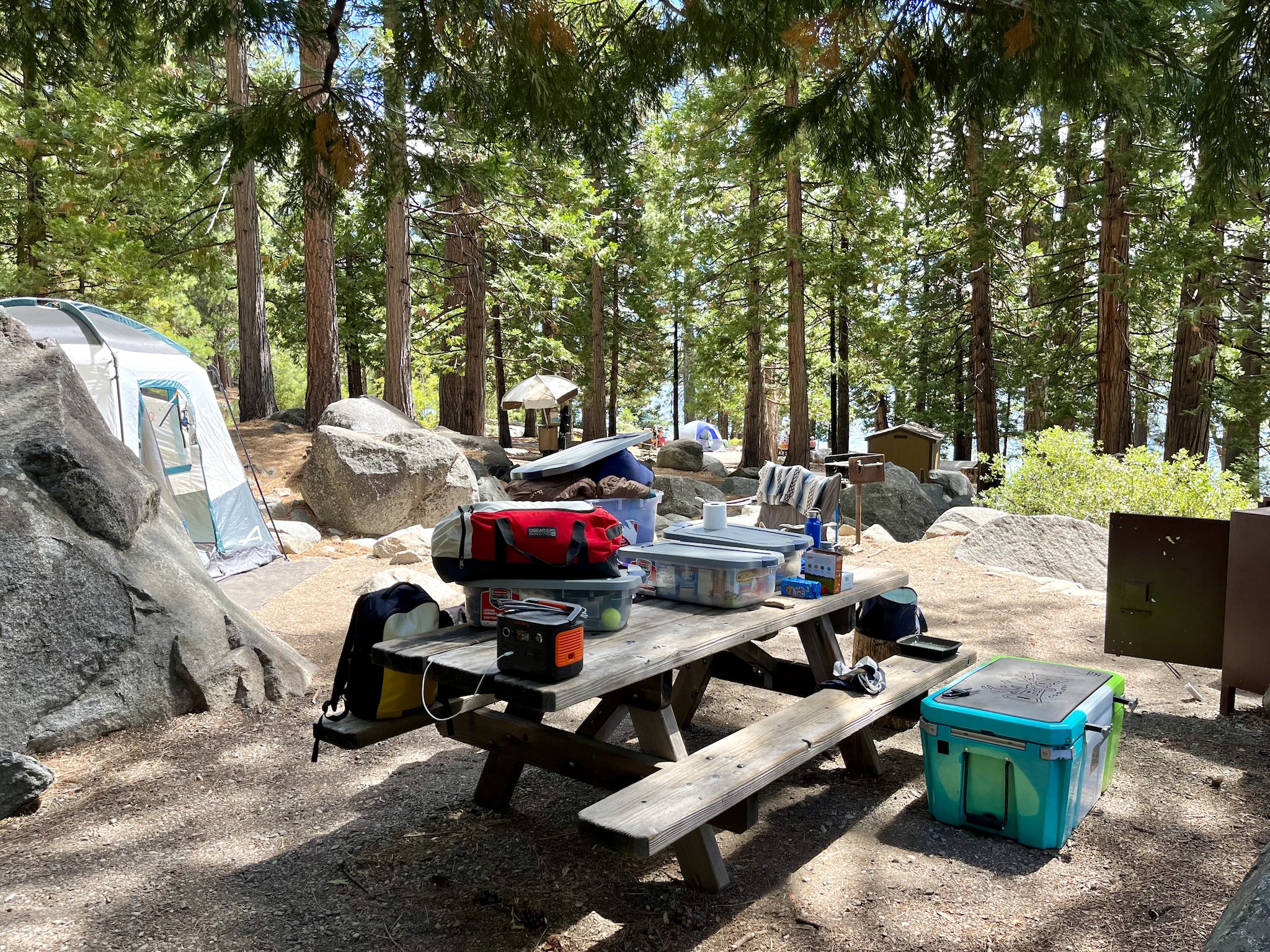 Campground, with a tent, fire pit, picnic table covered in boxes and duffle bags, a cooler, and a half-open bear box.