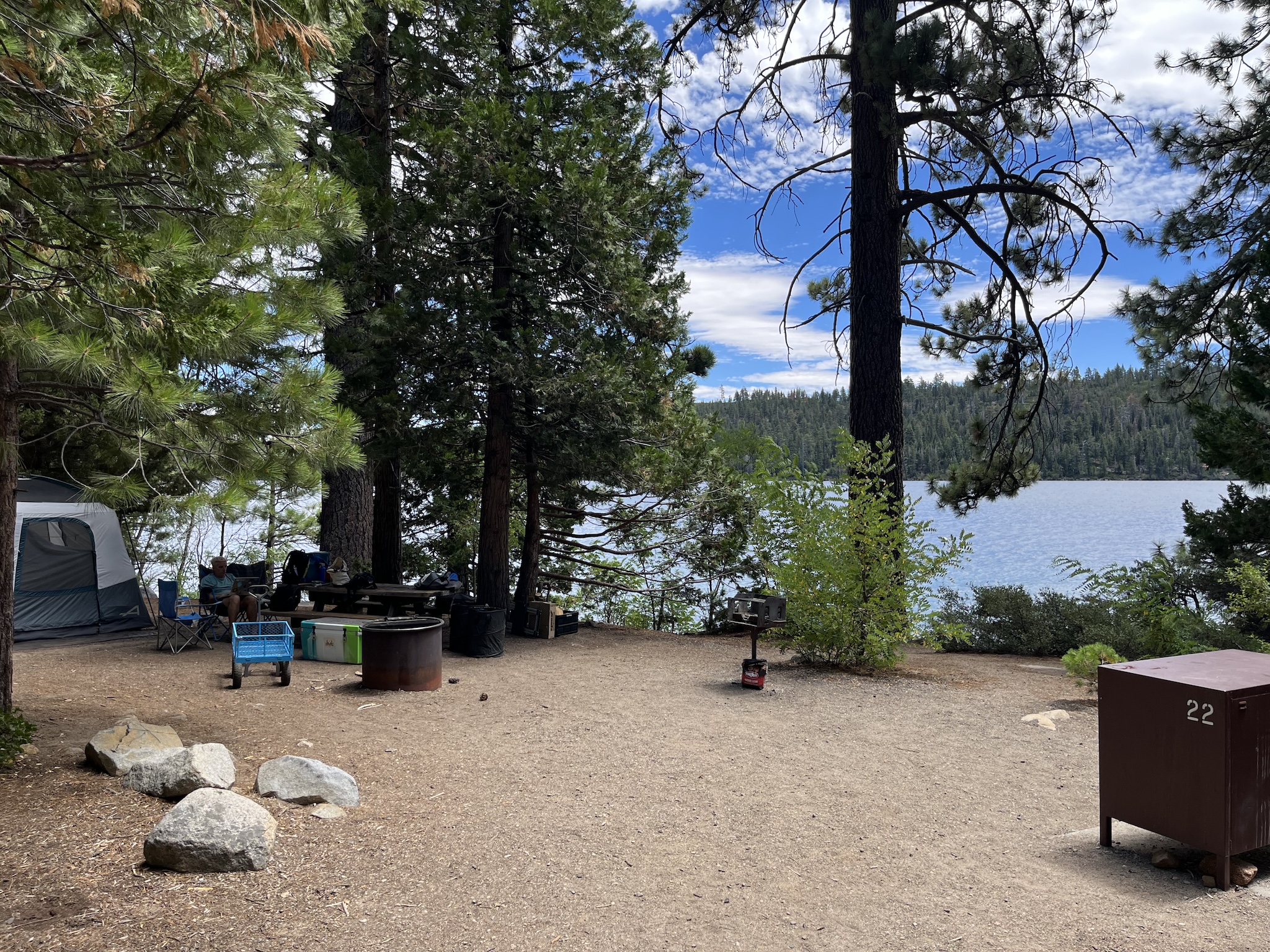 Campsite with a tent and picnic table in the background under some trees, lots of bare earth, and a bear box off to the side. In the background is a lake, right next to the edge of the campsite.