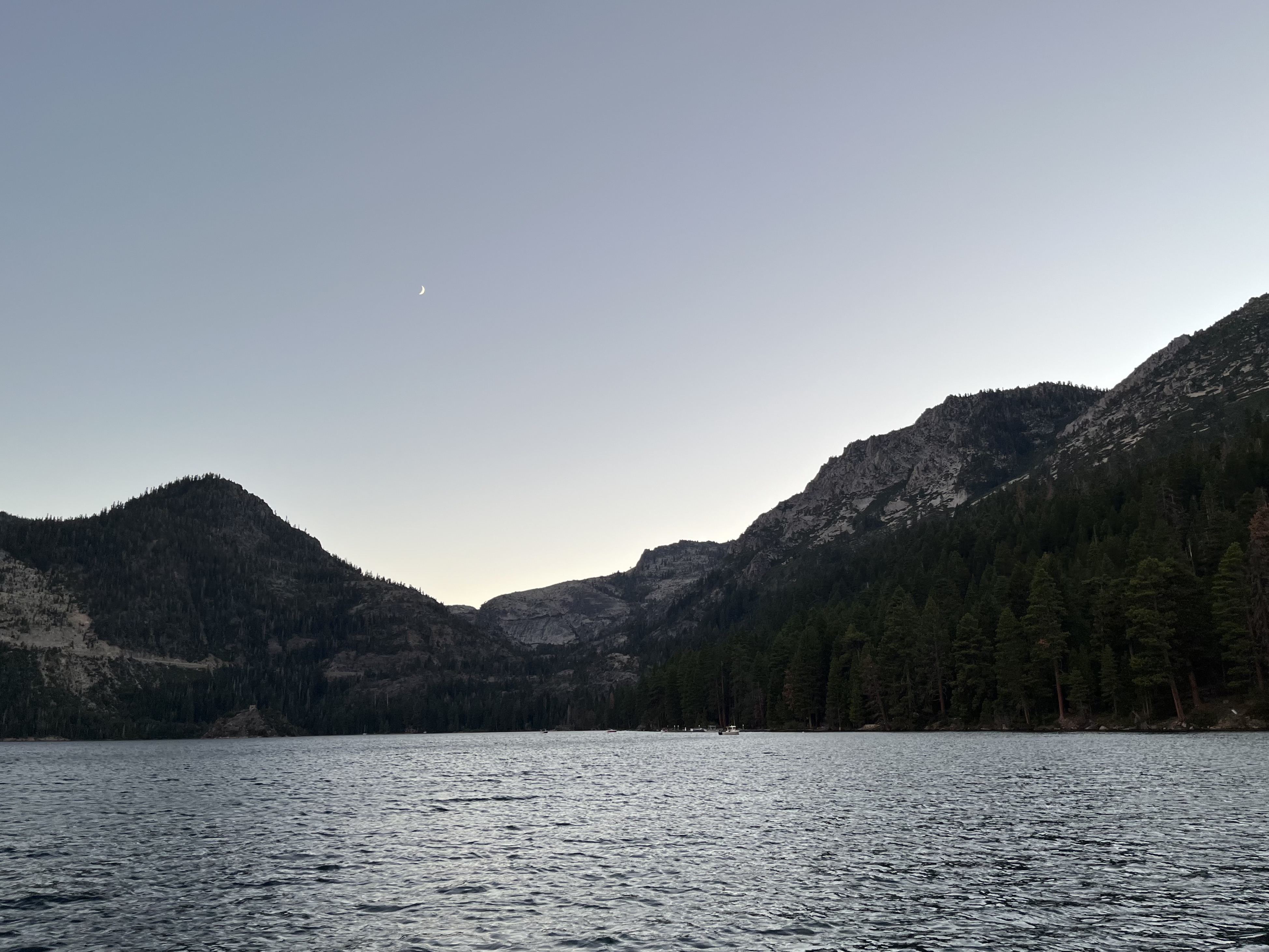 Two mountain peaks convering near the center of the photo, with a large expanse of lake in front. A sliver of moon is barely visible in the sky.