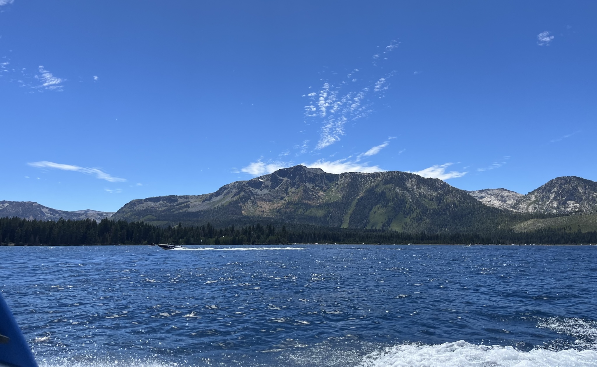 A large mountain with swaths of dark green pine trees and lighter green vegetation in the background. A deep blue lake with a speed boat jetting across it in the foreground.