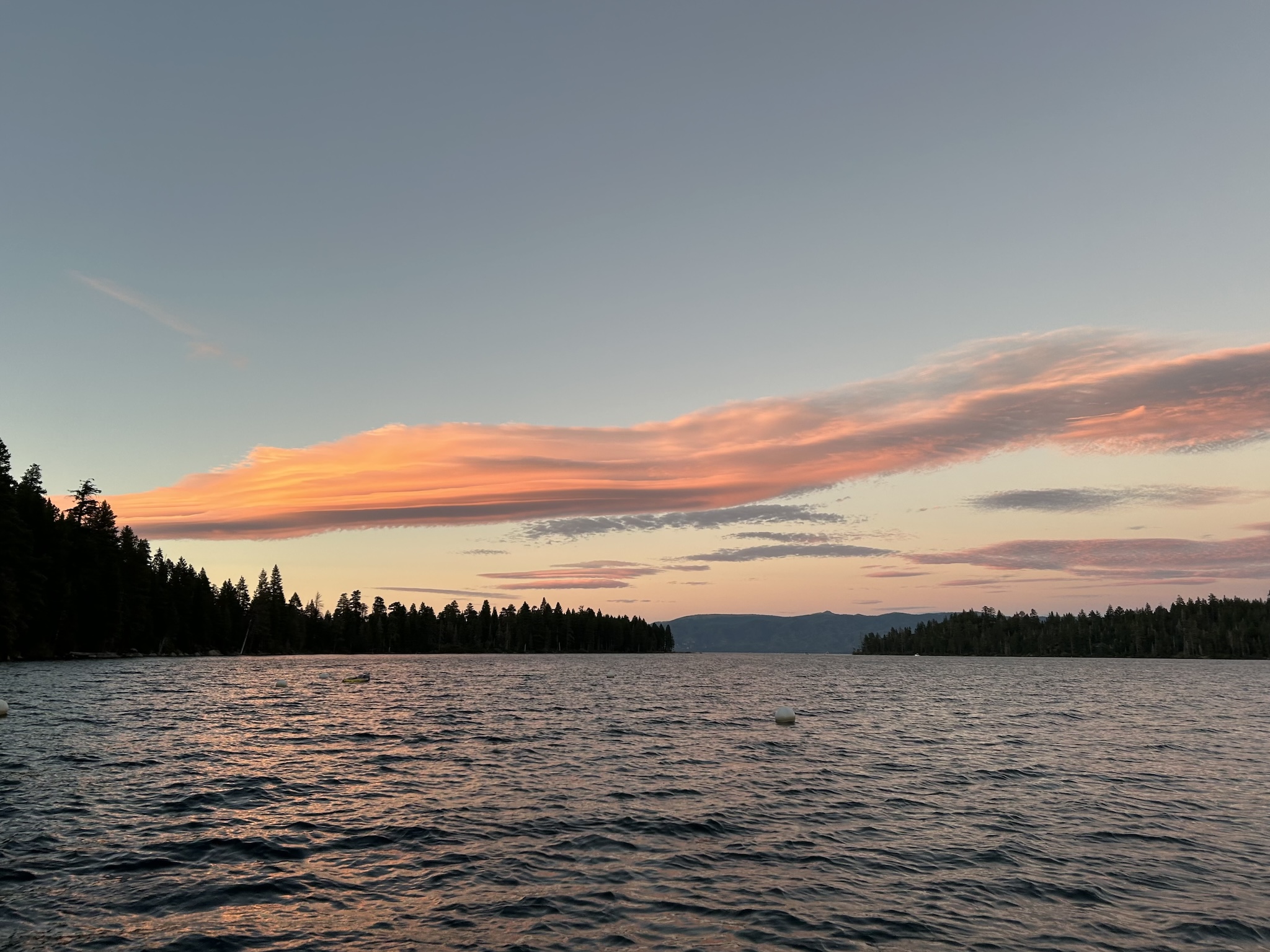 A large, lenticular cloud being lit in warm tones as the sun sets, hanging over the silluets of pine trees and the dark waters of a lake.