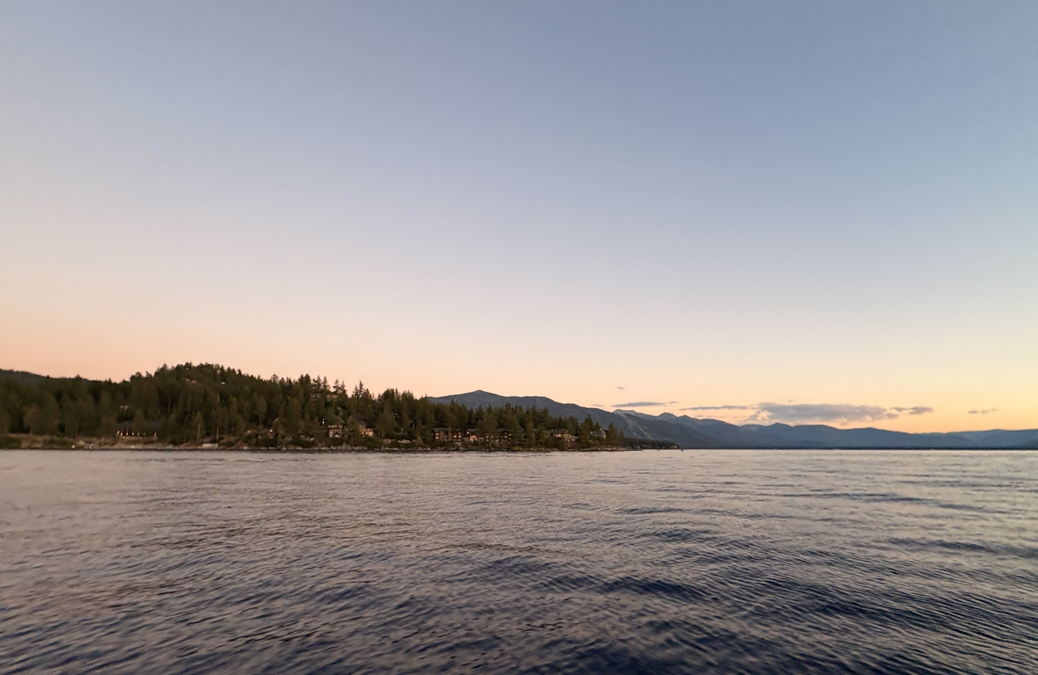 The shoreline of a lake, dotted with trees and a few houses. A ridge of mountains are visible in the background.