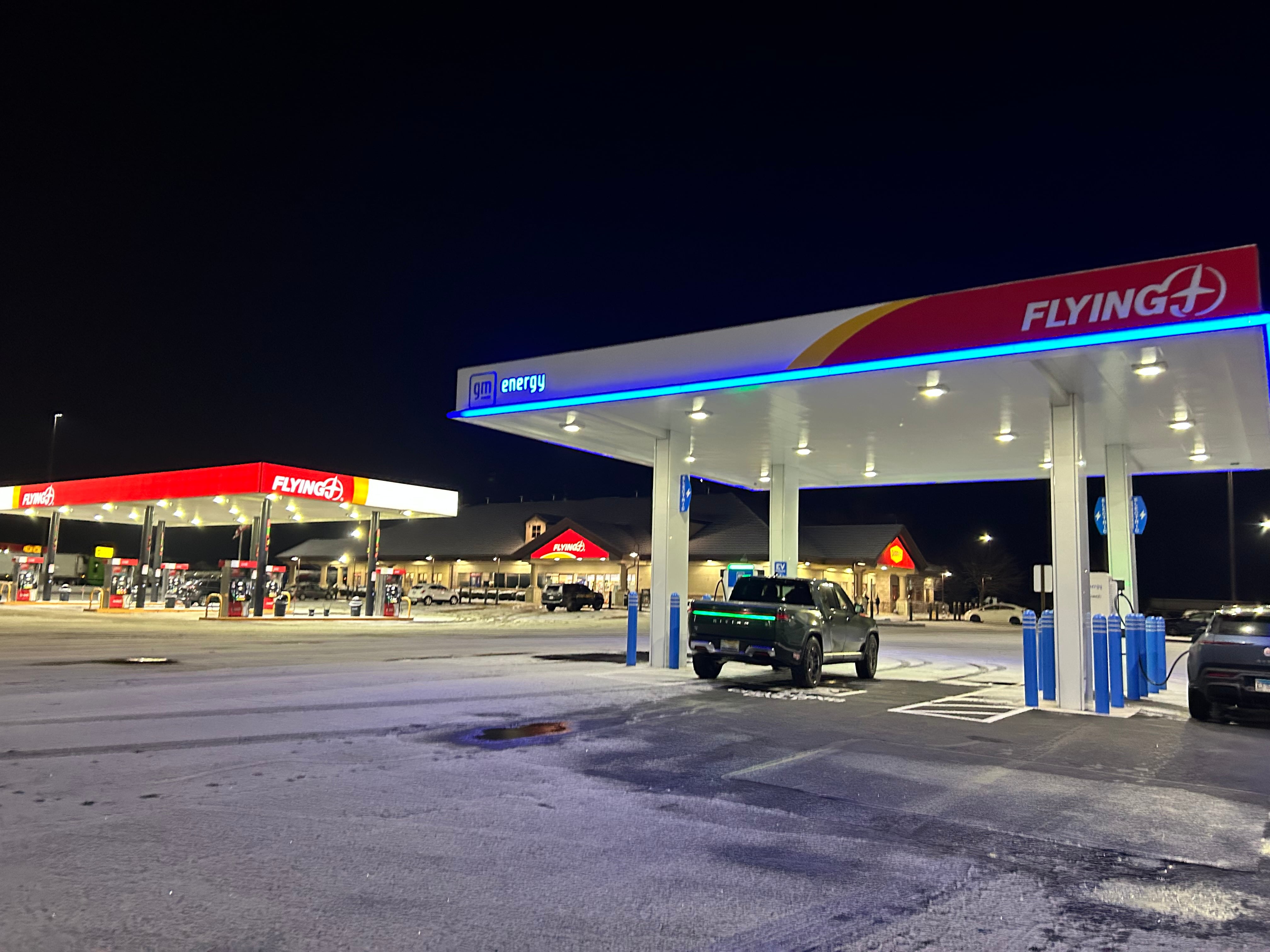 A truck parked under what looks like a standard gas station pavilion, but where the pumps are EV chargers. Regular gas station pumps and a gas station building are in the background.