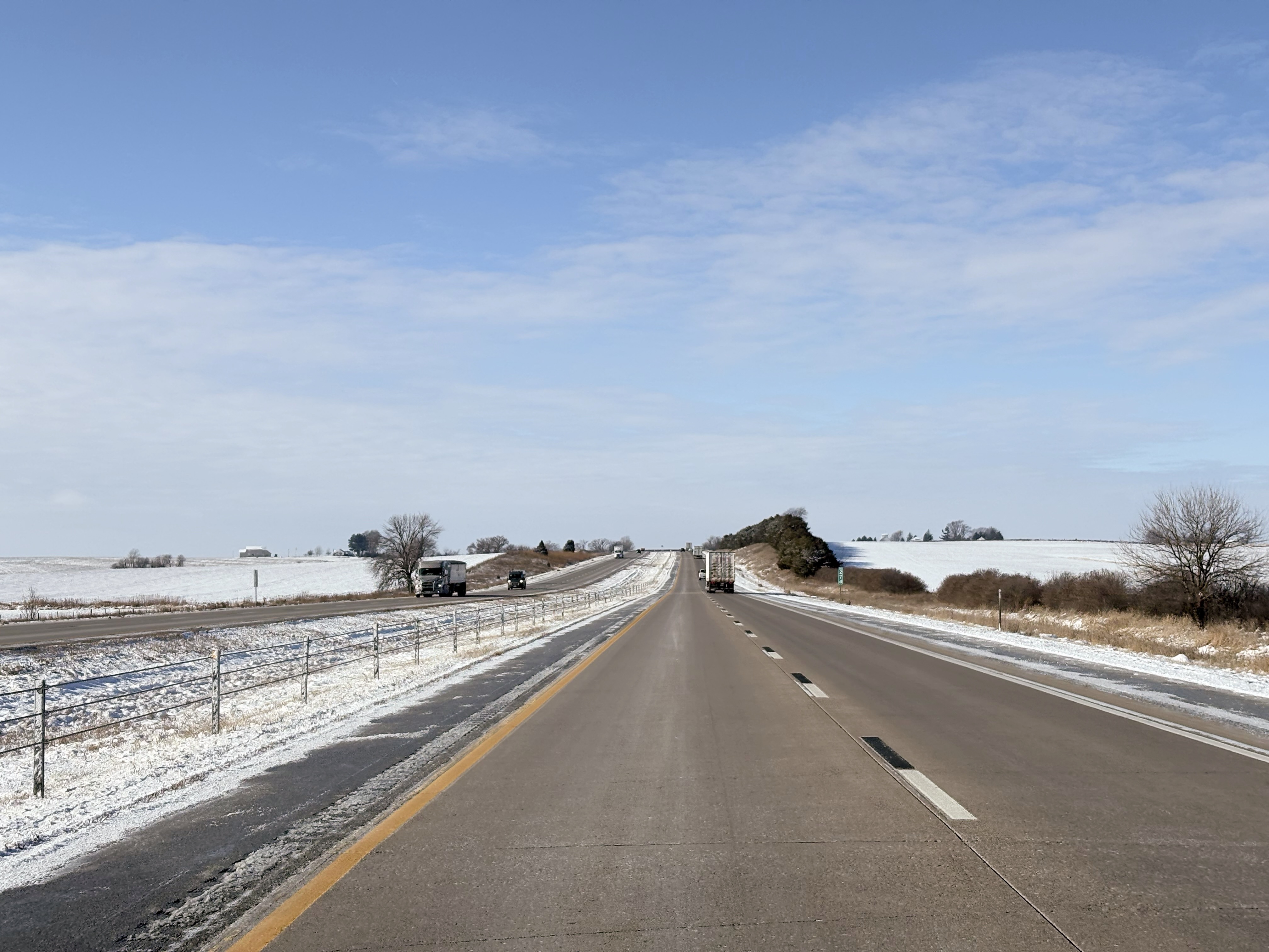 A stretch of highway arcing up into a small hill. The sides of the road are covered in snow and low plants.