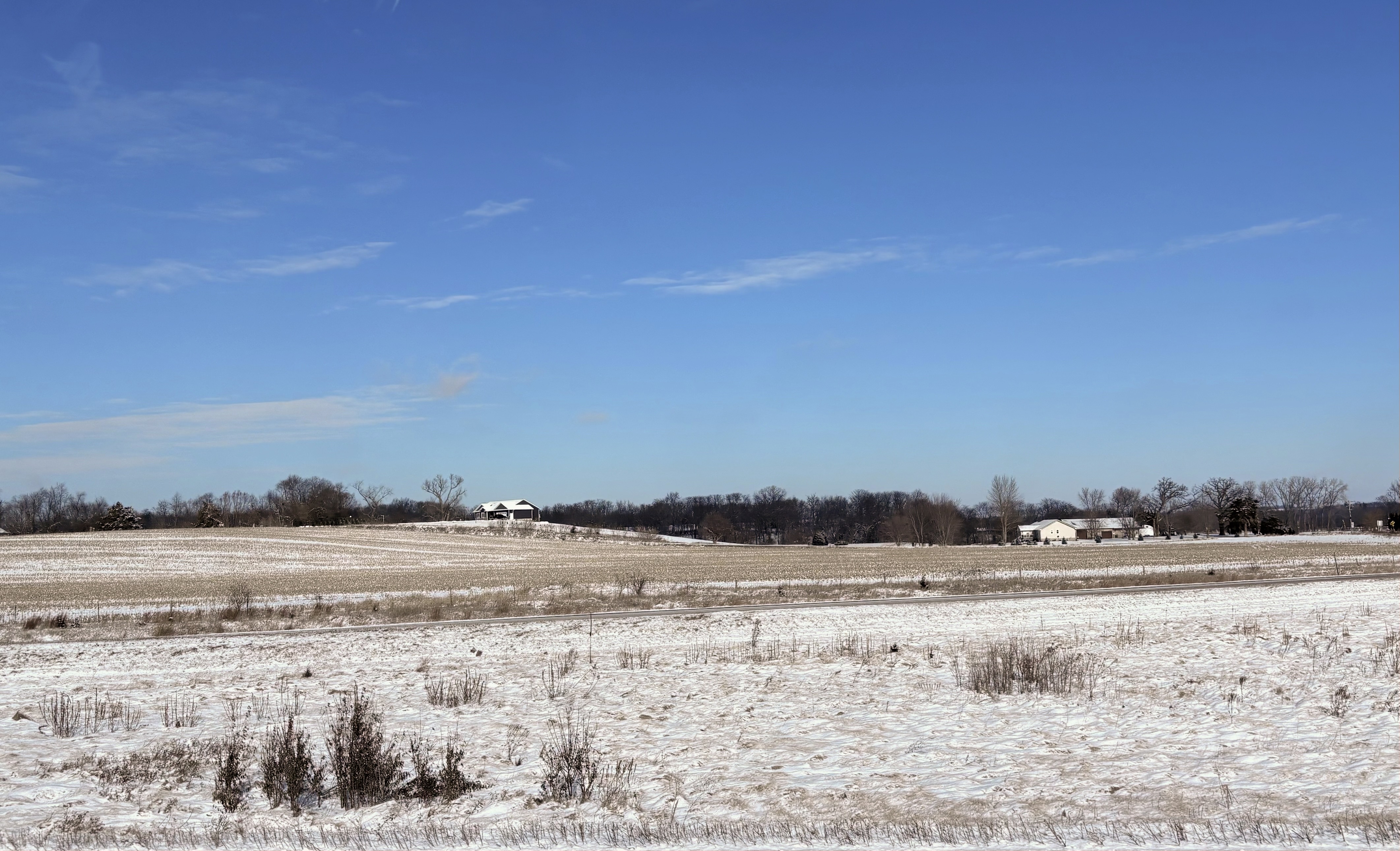 Two farmhouses sit in the distance between bare trees and against a blue sky with whisps of clouds. In the foreground are rolling hills with browned plants poking through snow.