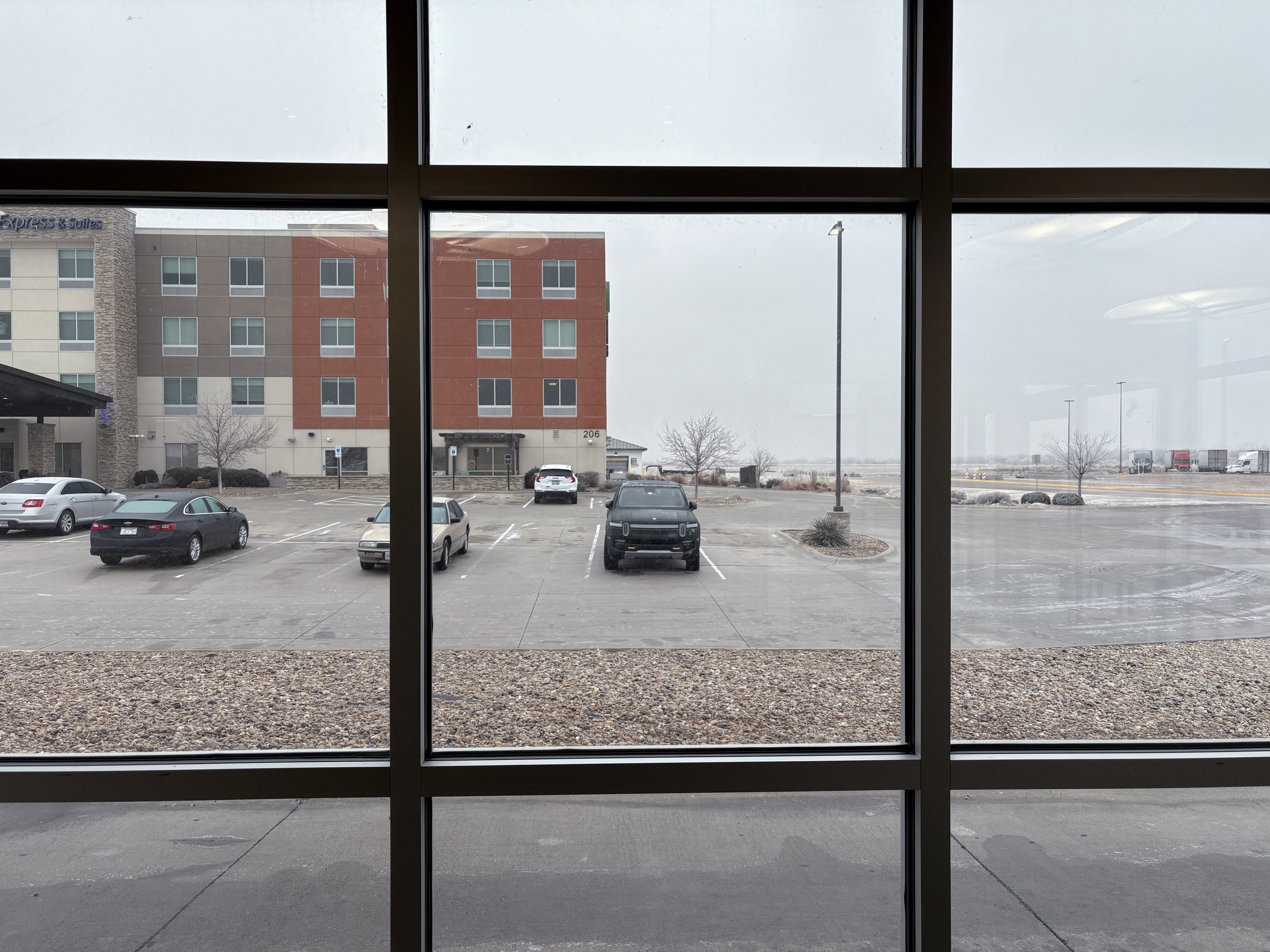 Hotel parking lot with several cars. The hotel is made up of several colored segements, and fog obscures anything in the distance. The photo is show from inside a building, and framed by a grid of windowpanes.