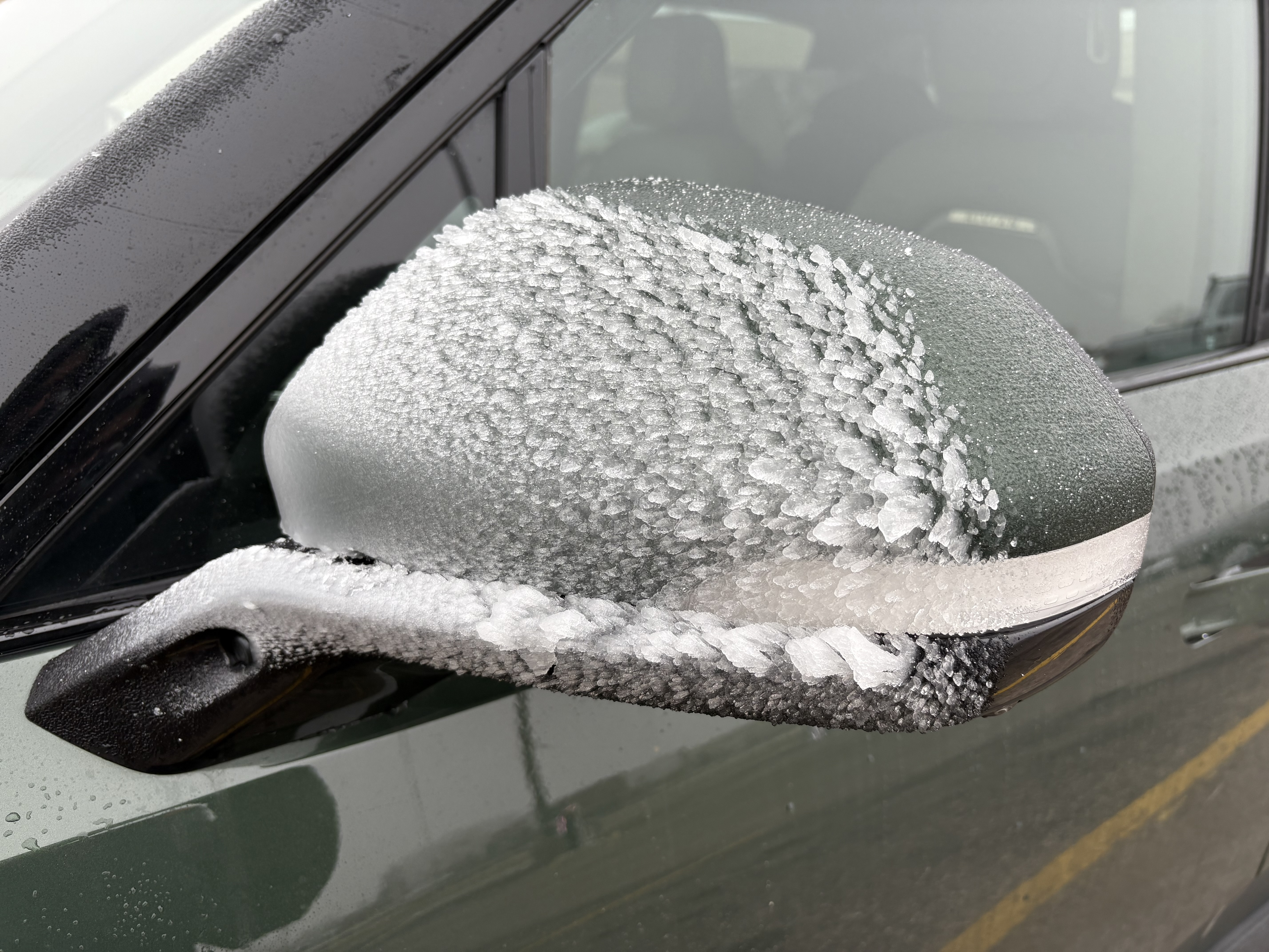 The driver side mirror of a truck, with ice frozen to the front in a strange formation of peaks that appear similar to reptile scales or small overlapping feathers.