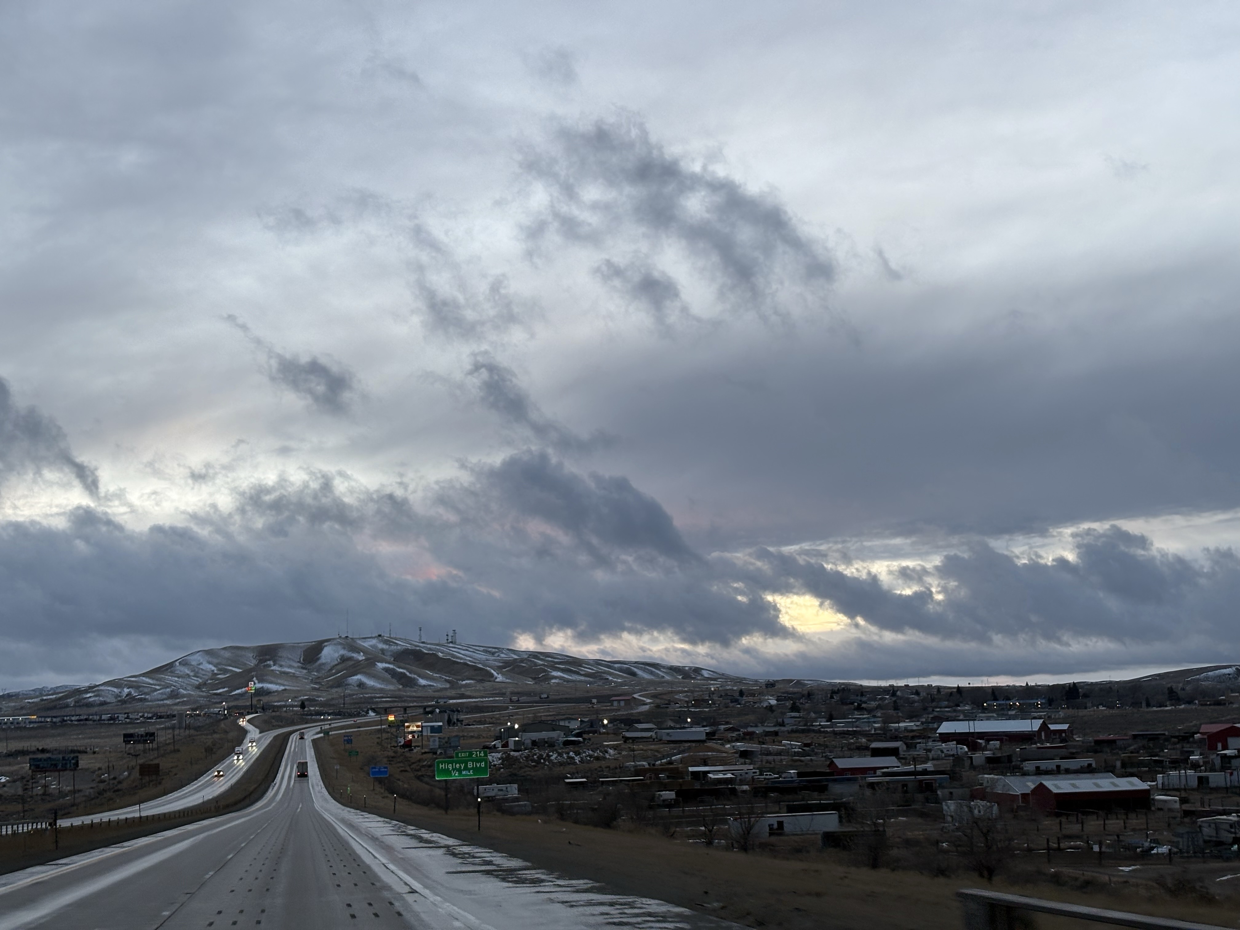 Wide angle shot of a low lying mountain with clusters of houses and a highway in the foreground. Clouds are dramatically lit by the setting sun