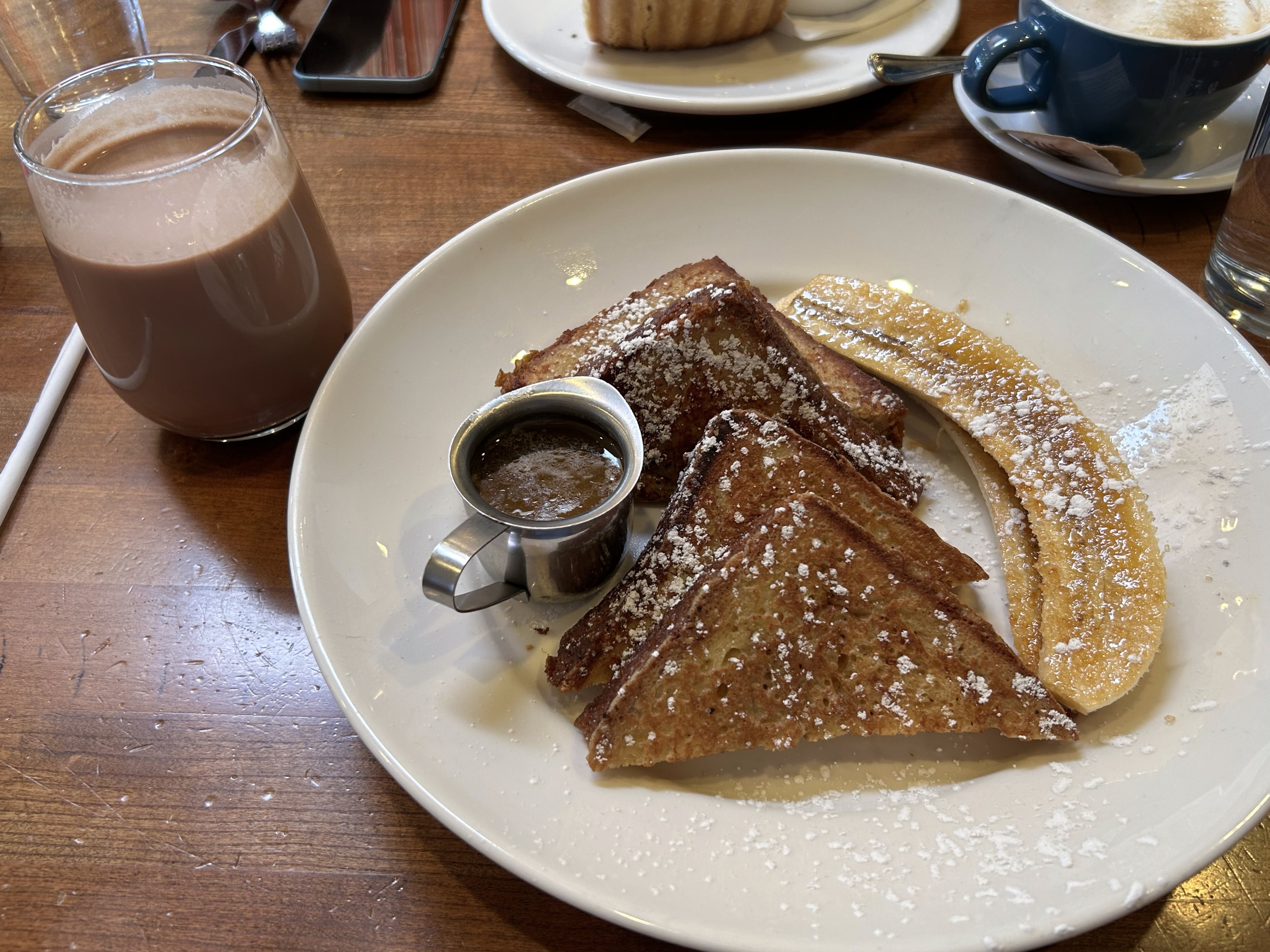 Plate with four slices of french toast, dusted in powered sugar, two slices of banana, and a small carafe of syrup. Glass cup filled with choolate milk.