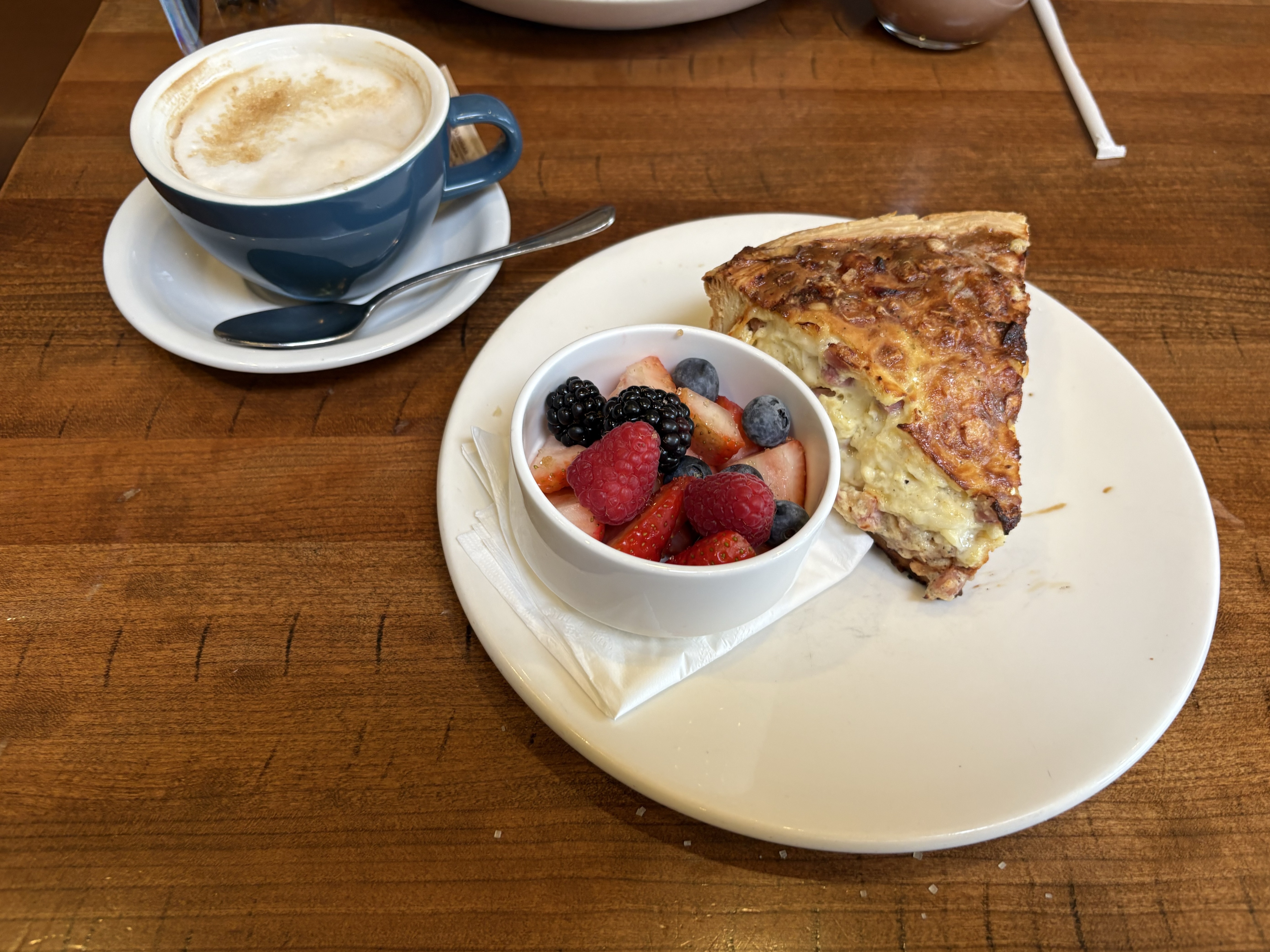 Plate with a slice of quiche and a small cup of strawberries, blackberries, and blueberries. Coffee cup containing a cappucino sitting on a saucer.