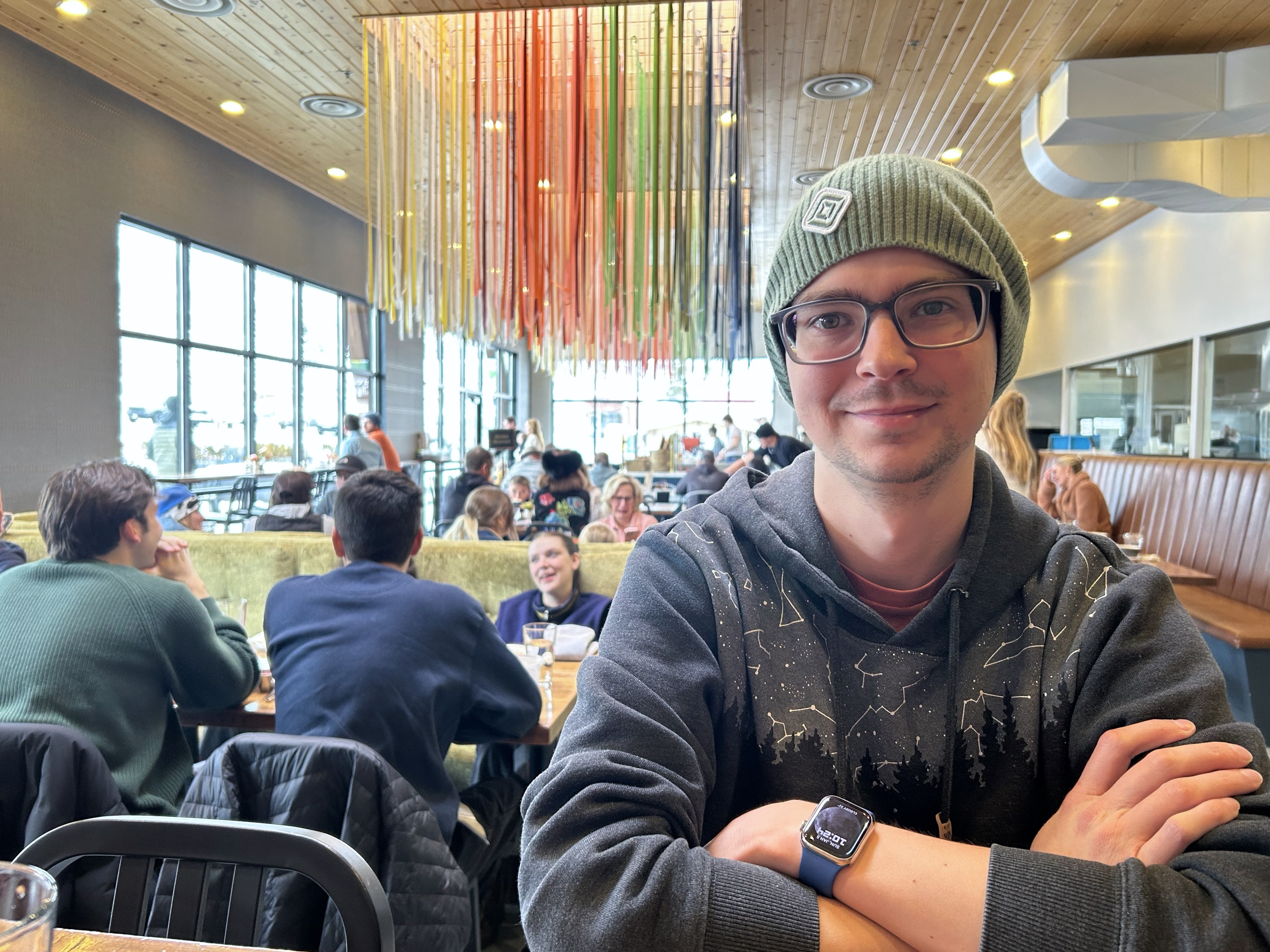 Person in a restaurant, wearing a green hat with a Rivian logo. Other patrons sit at tables in the background, and from the ceiling hangs a class art installation composed of numerous colored glass or plastic tubes.