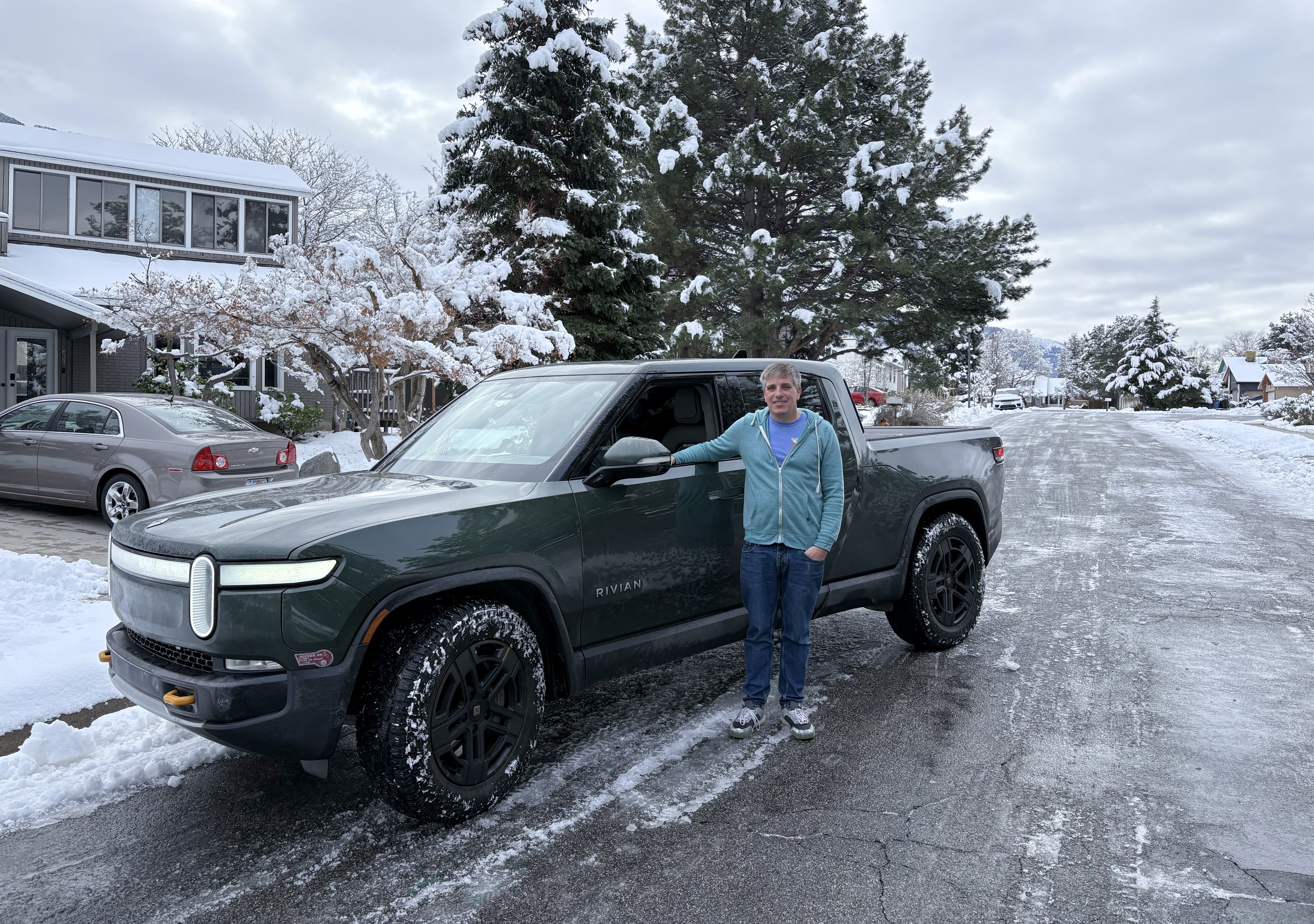 Person standing in front of a green truck. The truck is parked on an icy street with houses and trees behind it.