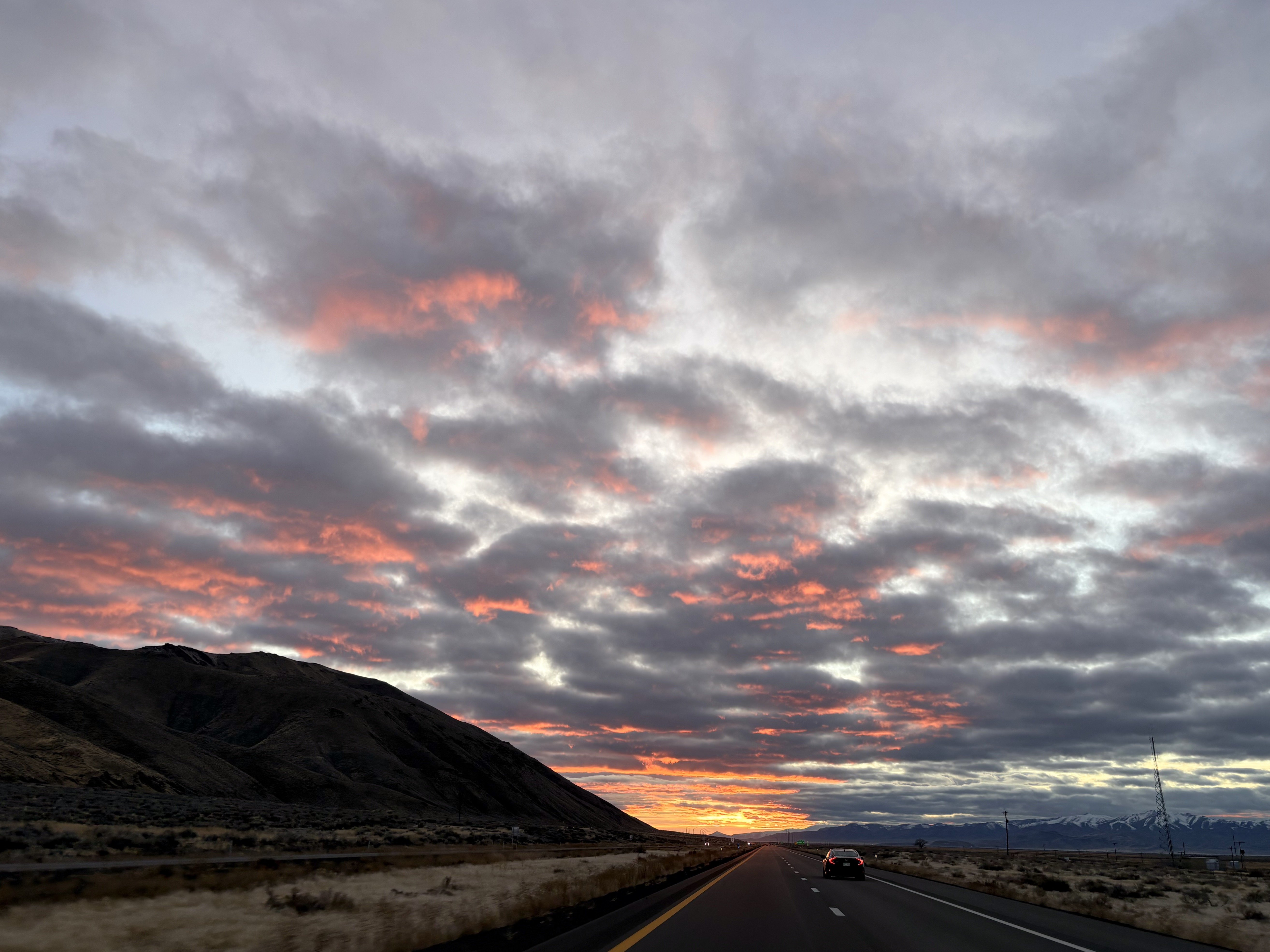 Stretch of highway with a small hill on the left and plains on the right. A partly cloudy sky is lit by the sun, showing bright orange highlights on the underside of the clouds.