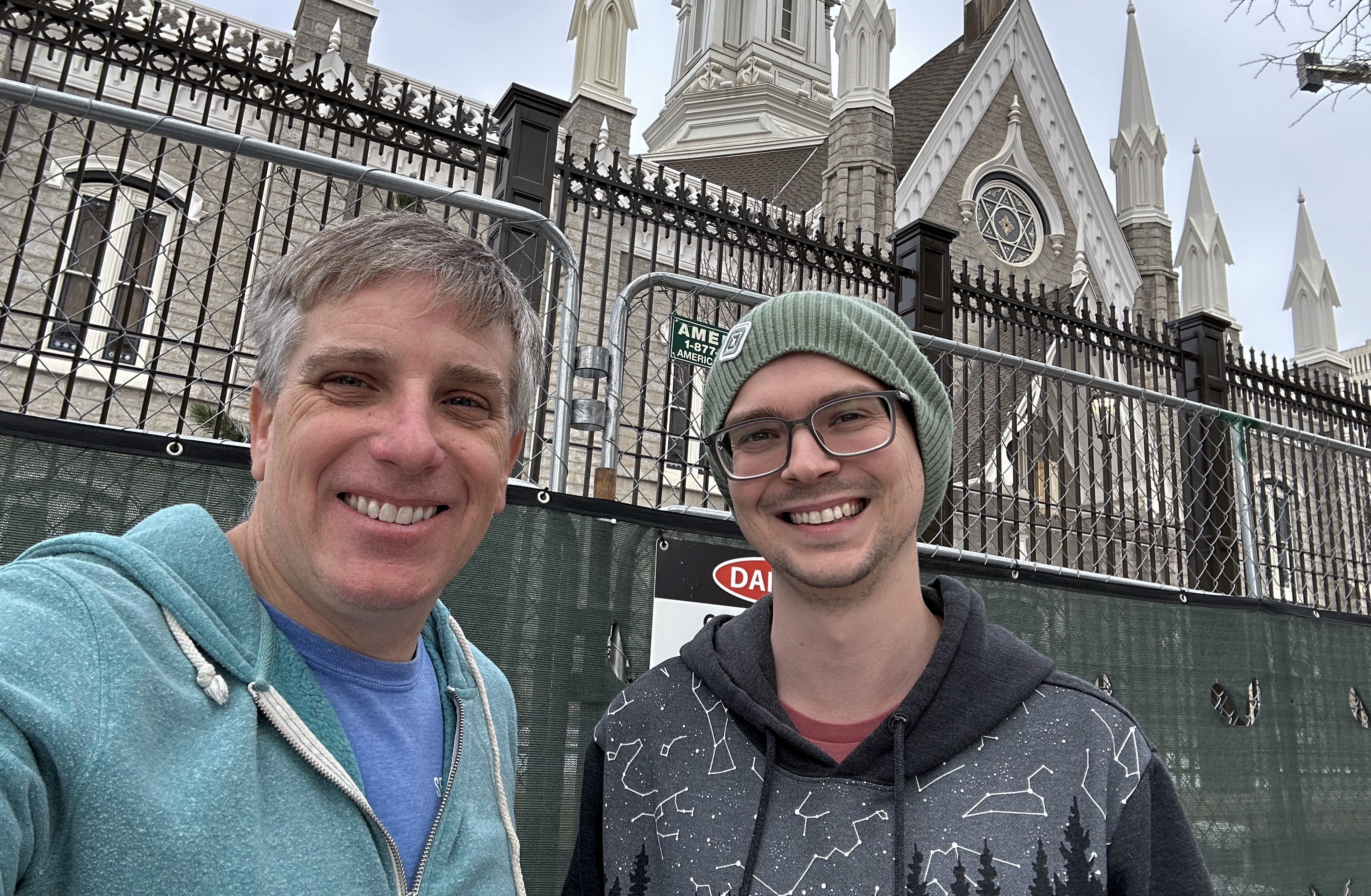 Two people pose for a selfie in front of a construction fence with a danger sign. An ornate church-like behilding is behind the fence.