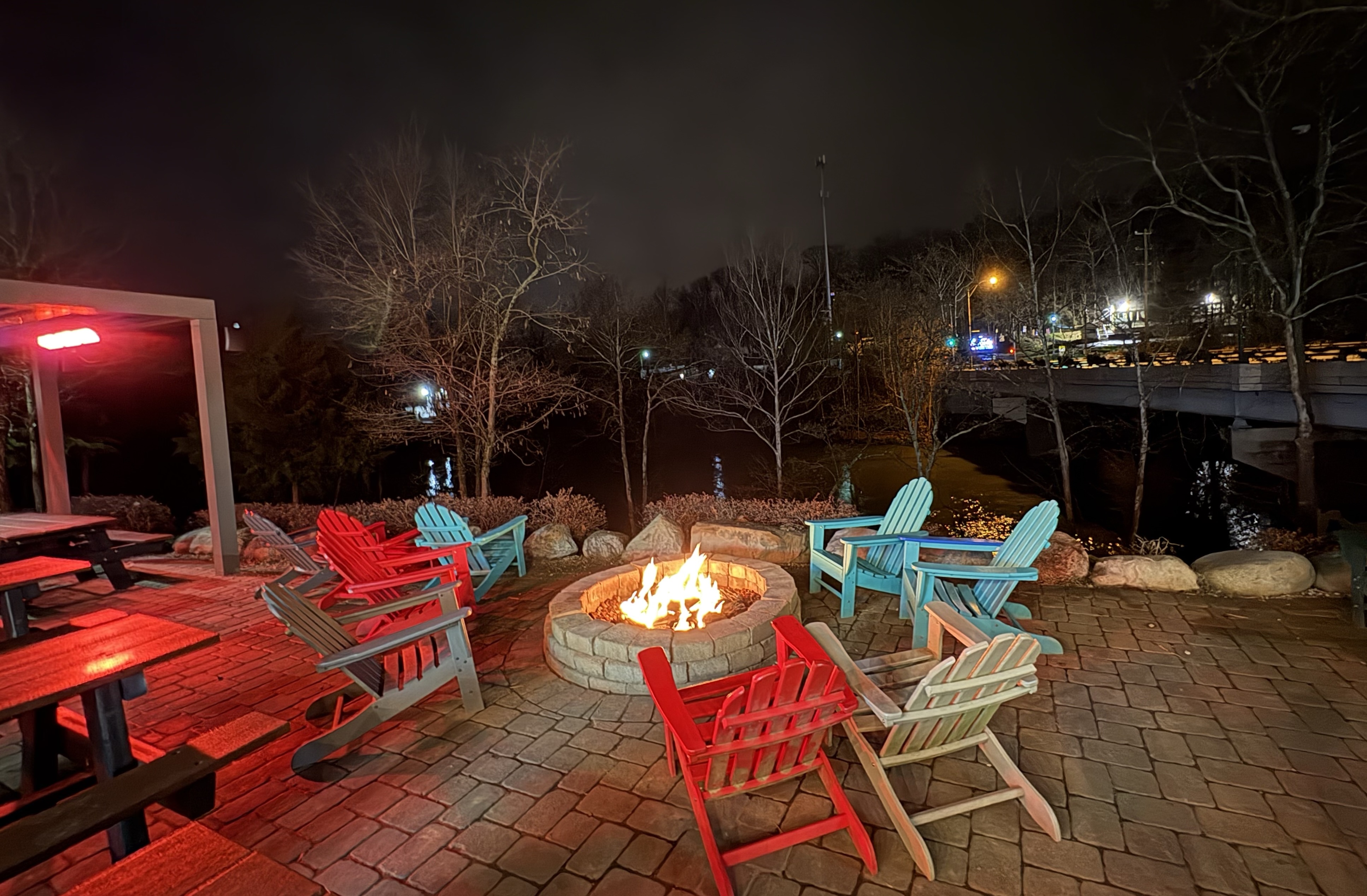 Photo at night time. Several adarondak style chairs around a lit, brickfire pit. A dark river and bridge are visible in the background.