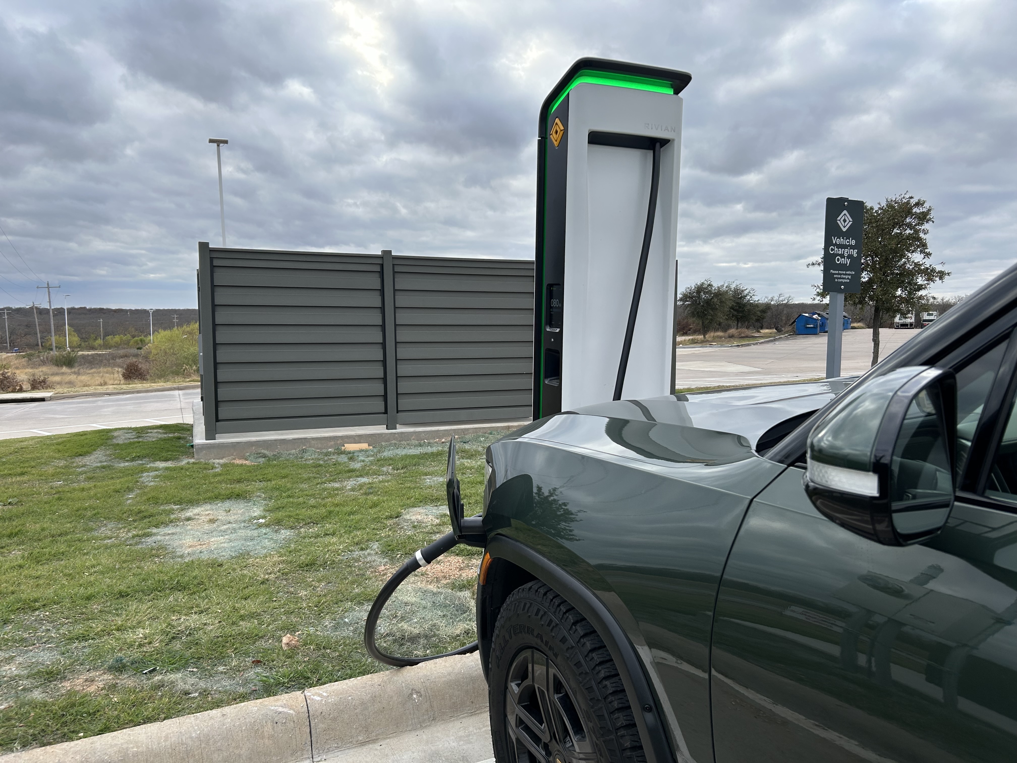 Tightly cropped photo of the front of a truck, plugged in to a Rivian charging station. In the background the sky is cloudy, and there is a small strip of grass behind the curb of the parking lot.