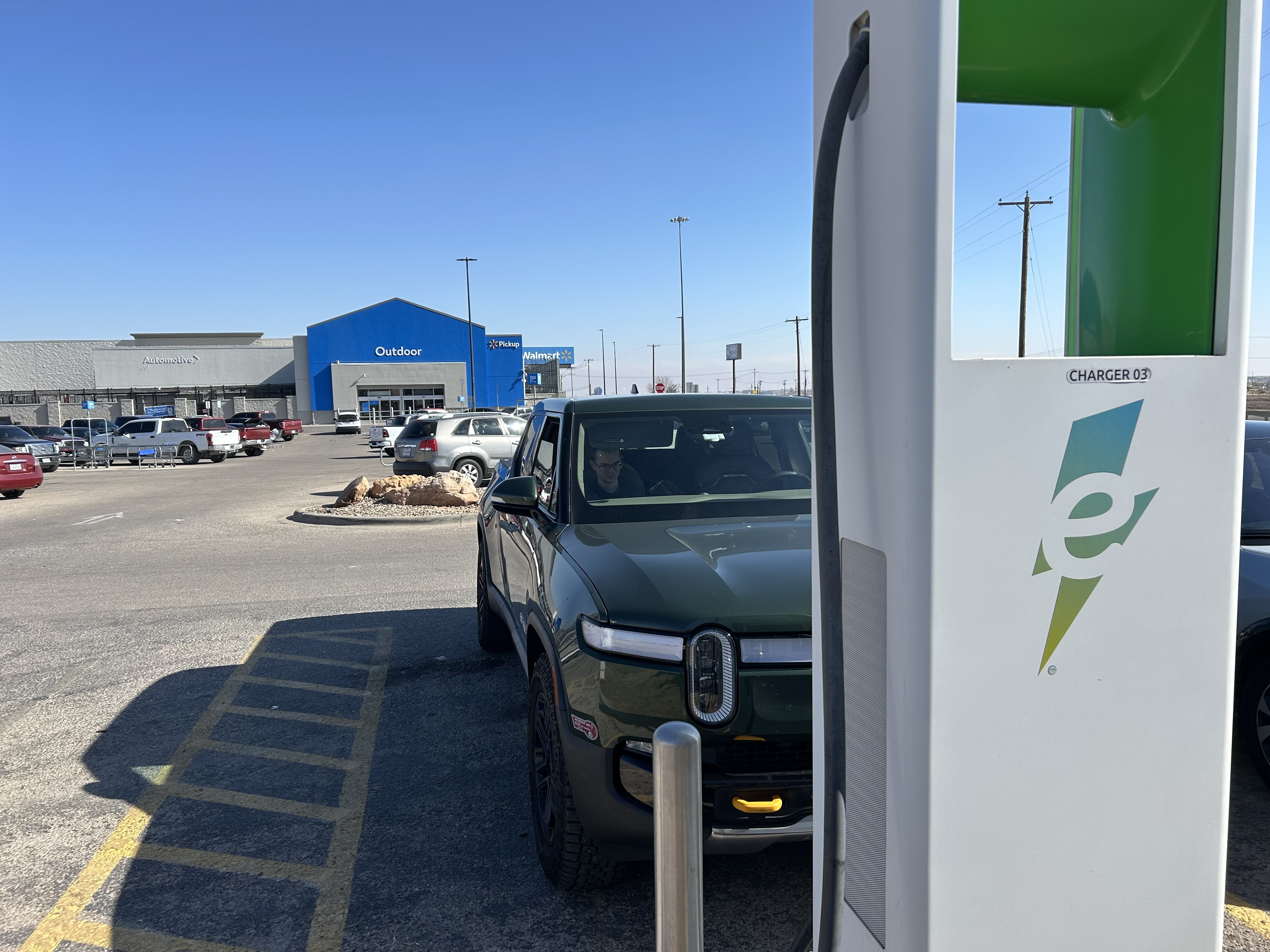 Photo of an Electrify America charging station, with a truck plugged in behind it. A Walmart parking lot and store are in the background of the photo.