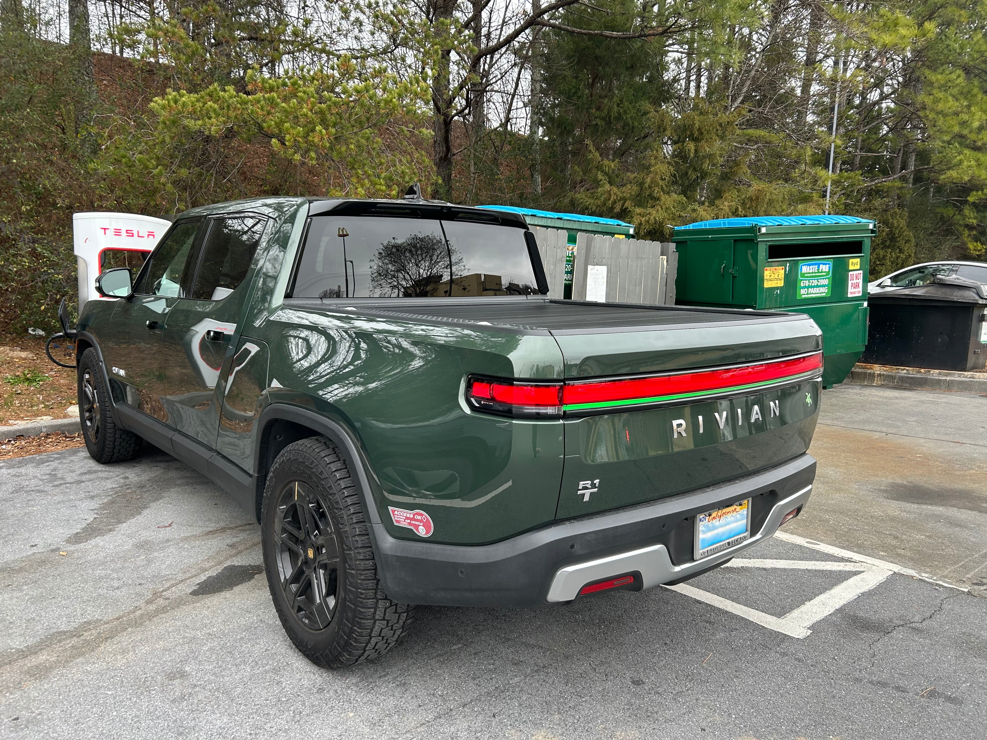 A Rivian truck in a parking spot. Behind the charging station are some large trash recepticles.