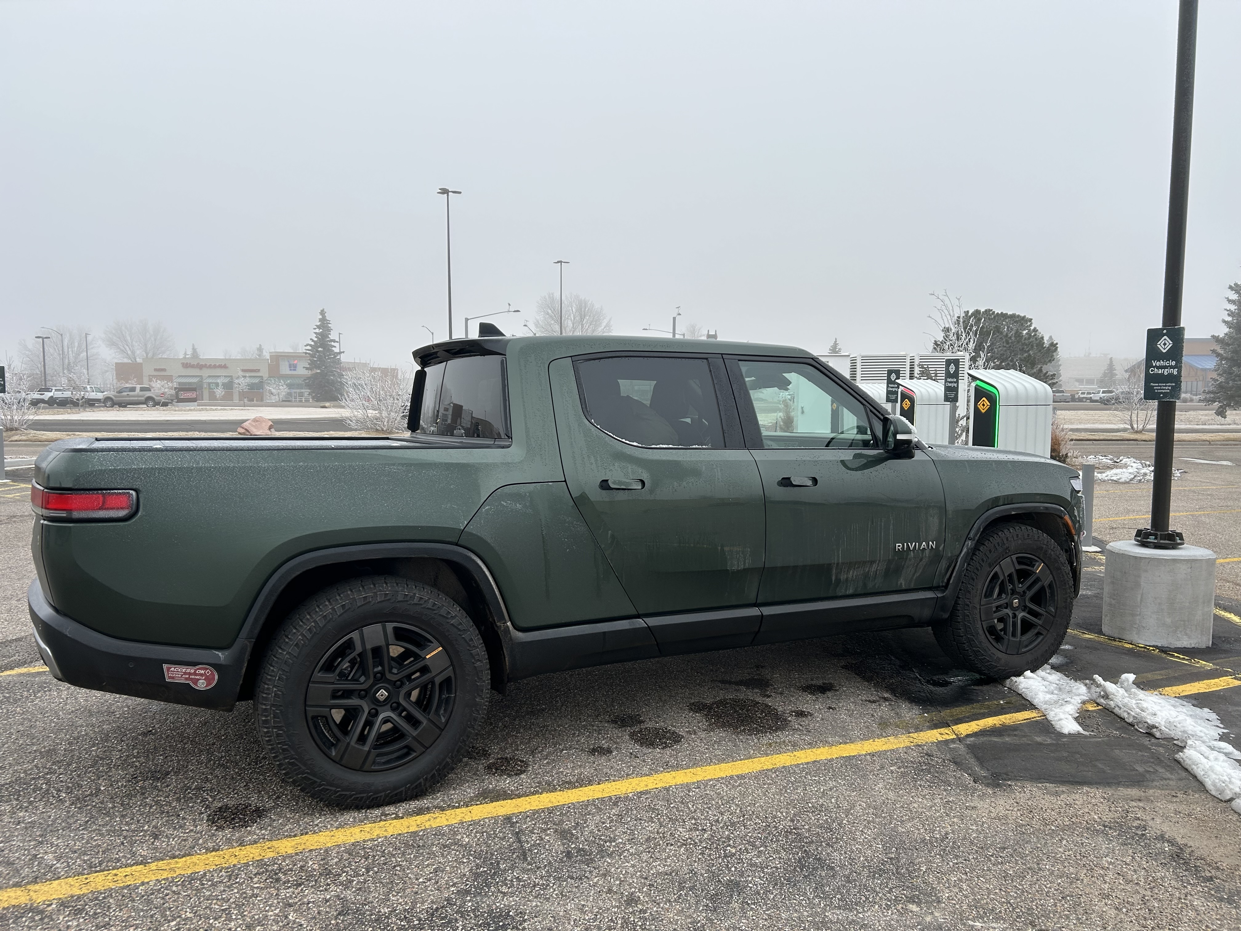 Rivian truck in a parking lot, with Rivian charging stations behind it. The sky is dark gray, and the background is very foggy, so you can’t see much.