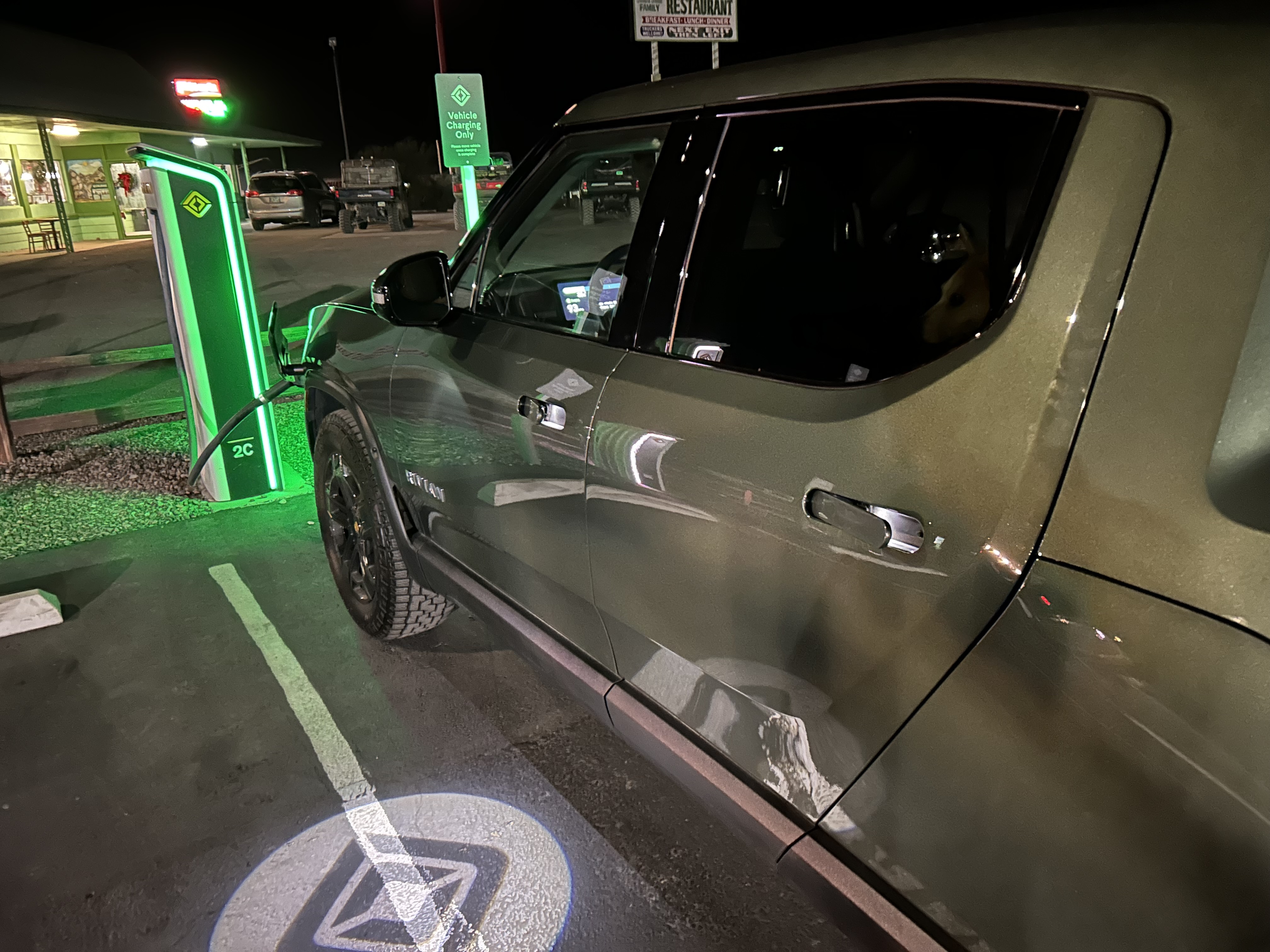 Close up photo of a truck plugged in to a Rivian charging station at night. There is a diner in the background, and beside the truck the Rivian logo is being projected on to the ground from the truck’s door handle.