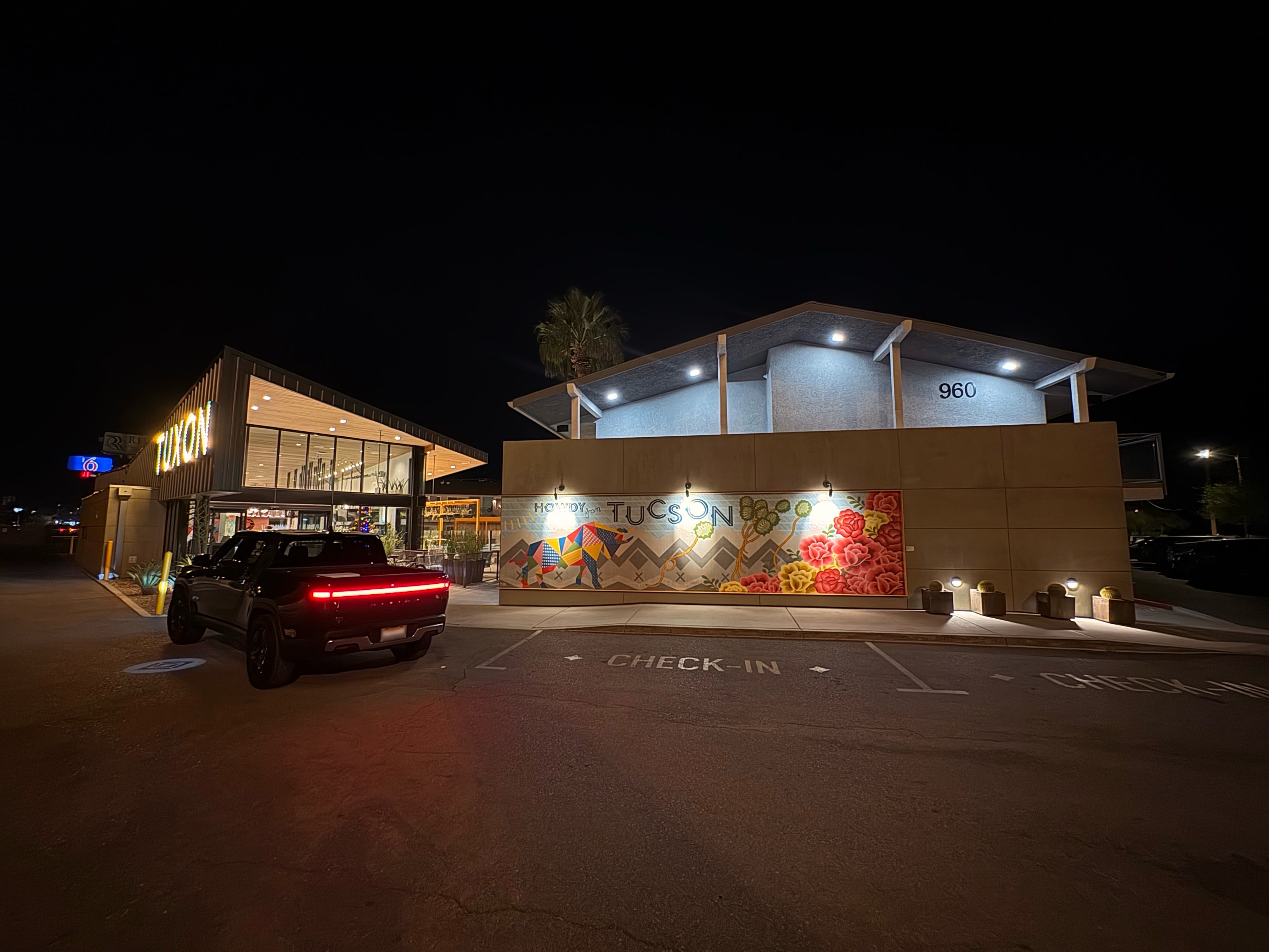Two buildings with a truck parked in front. One building has a light up sign reading “Tuxon” in all caps, and the other has a mural with illustraited mountains and flowers and the word “Tucson”.